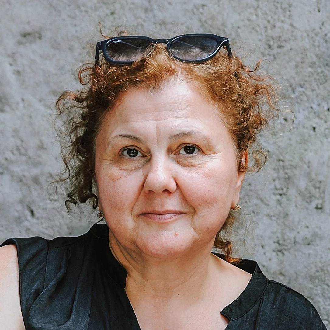 Nahaufnahme einer Frau mit roten, lockigen Haaren, Sonnenbrille auf dem Kopf, trägt eine schwarze Jacke, steht vor einer grauen Wand.