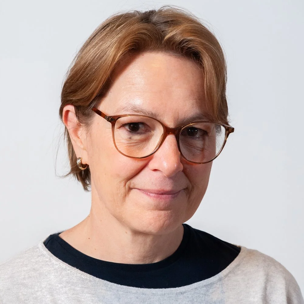 Porträt einer Frau mit kurz braunen Haaren, Brille und Ohrringen vor einem weißen Hintergrund.