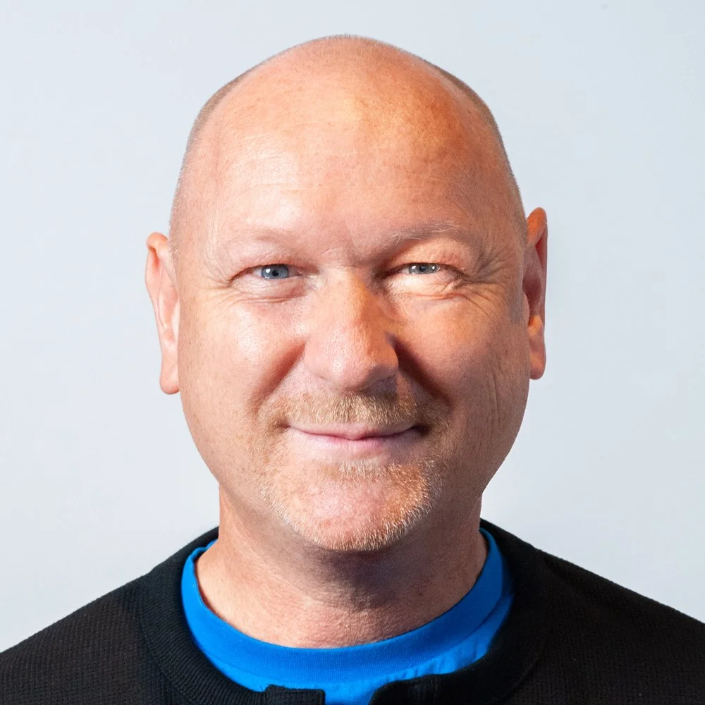 Nahaufnahme eines lachenden Mannes mit Glatze und blauen Augen, trägt blauen und schwarzen Kleidung vor hellem Hintergrund.