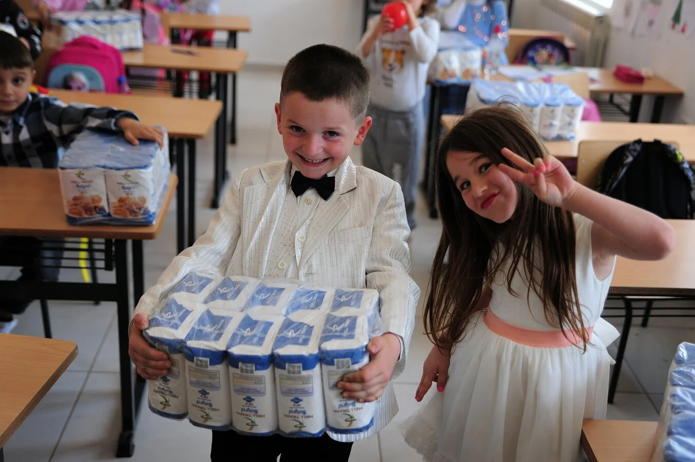 Zwei Kinder in der Klasse, ein Junge hält mehrere Packungen Mehl, eine Mädchen zeigt ein peace-Zeichen und lächelt in die Kamera.