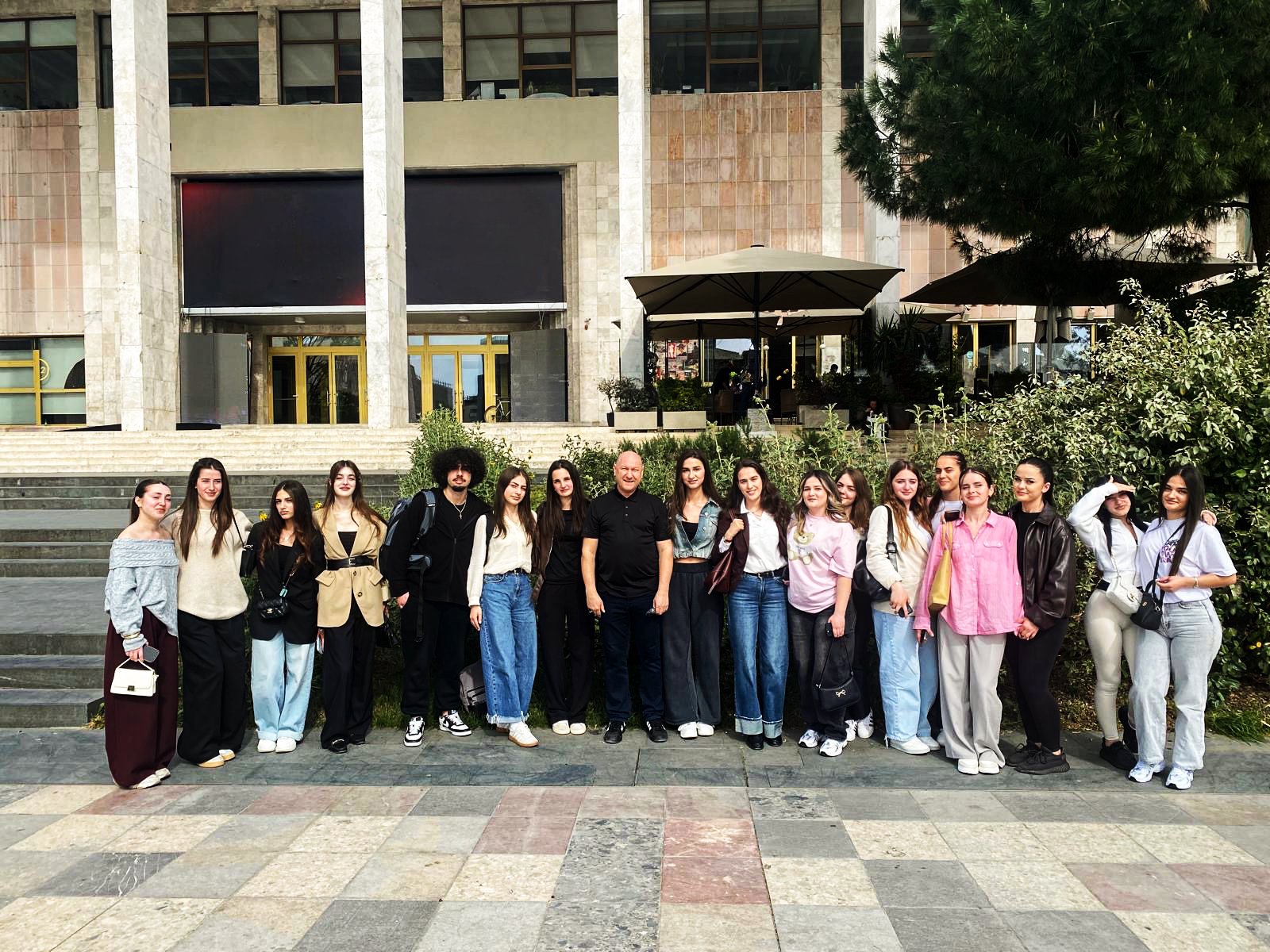 Eine Gruppe von circa 18 Studierenden steht vor einem modernen Gebäude mit Steinstufen und einer begrünten Umgebung, sie posieren für ein Gruppenfoto im Freien.