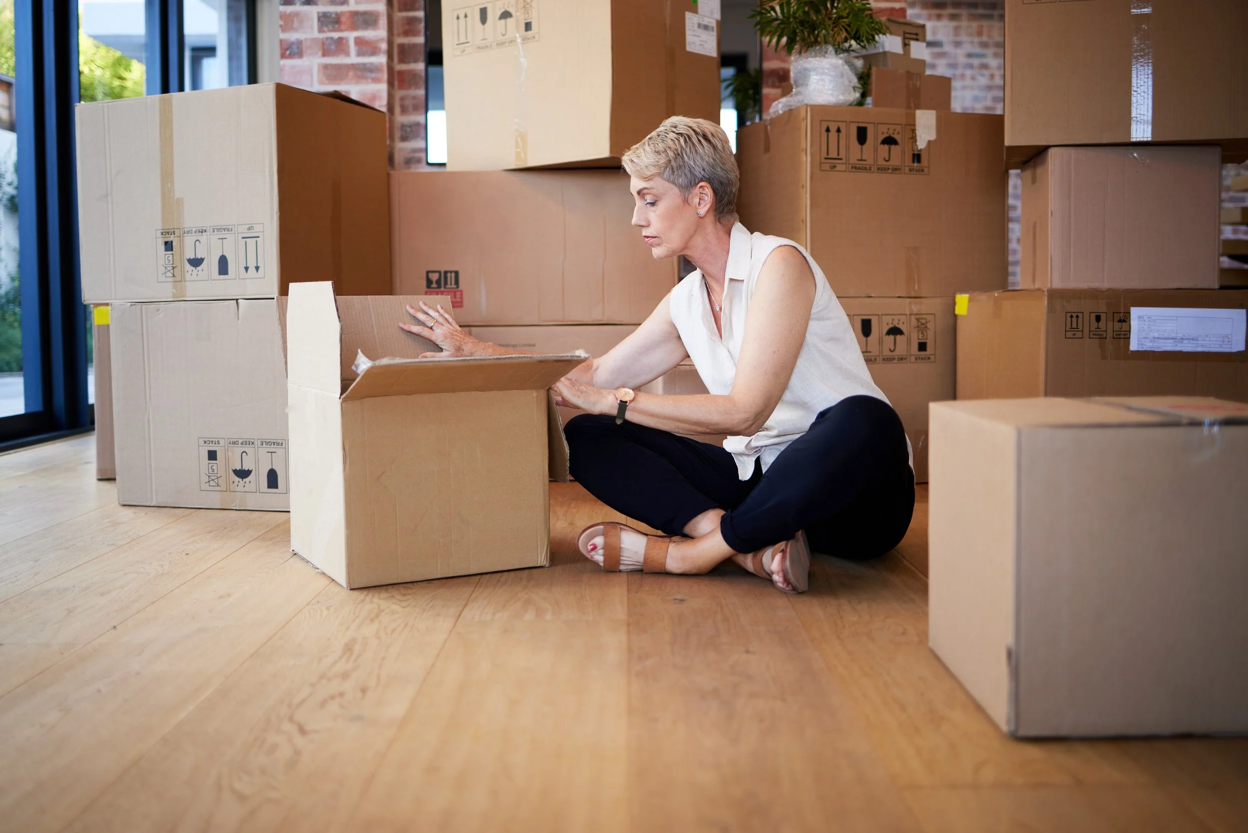 A woman with short gray hair, sitting on the wooden floor, opening a cardboard box in a room filled with stacked moving boxes.