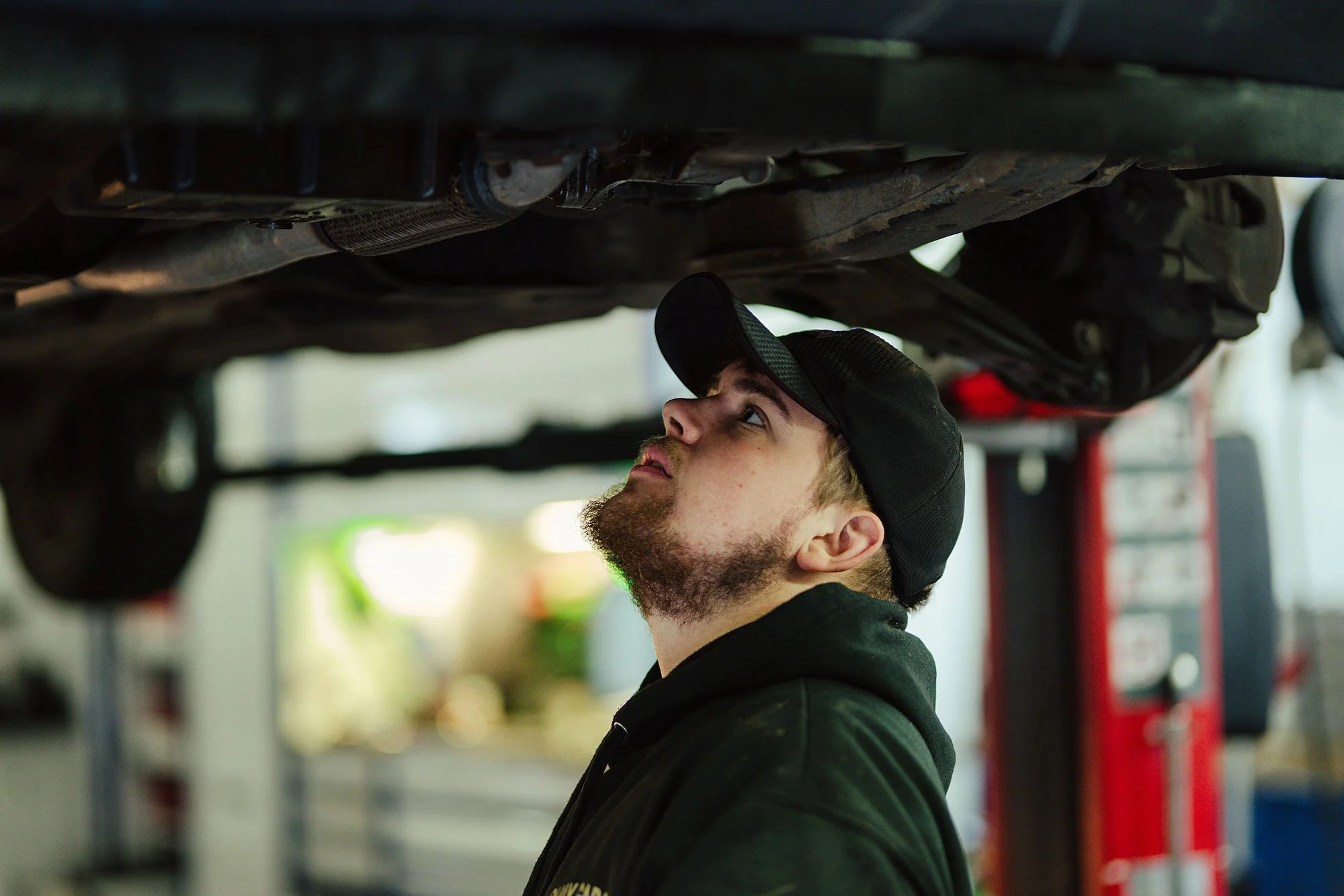 A man with a beard and black cap inspecting the underside of a vehicle in an auto repair shop.