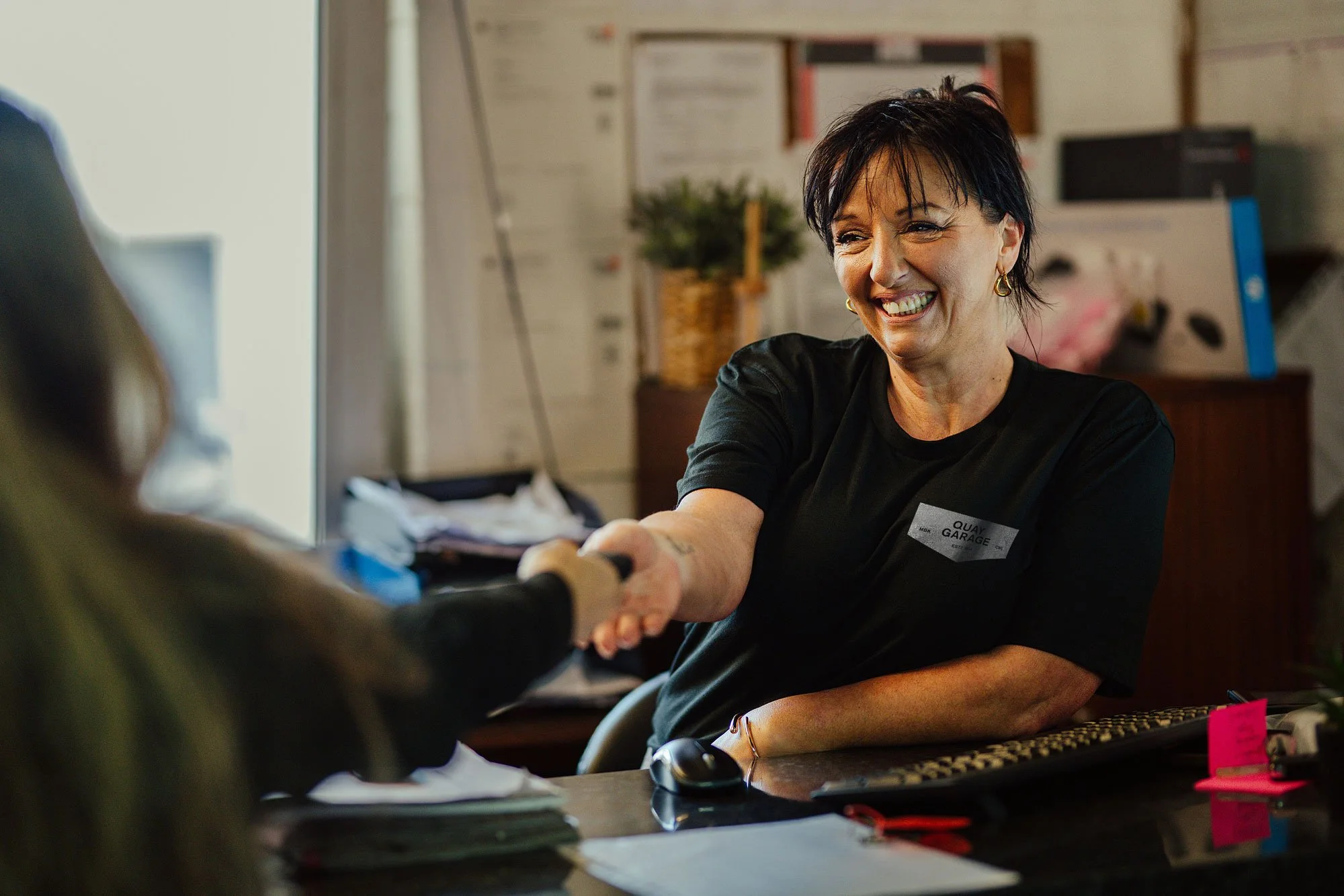 A woman shakes hands with a customer at a reception desk, smiling warmly. She is wearing a black t-shirt with a name tag that reads "QUAY GARAGE." The desk has papers, a keyboard, and pink sticky notes. The background includes a bulletin board, a plant, and office supplies.