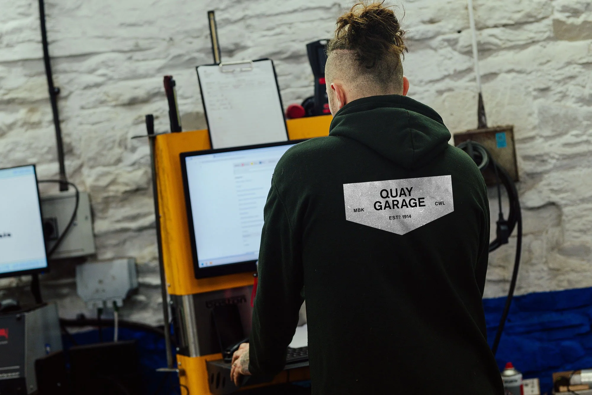 A person with curly hair and a shaved side, wearing a black hoodie with a "Quay Garage" graphic on the back, working on a computer in a workshop with a white brick wall, tools, and supplies around.