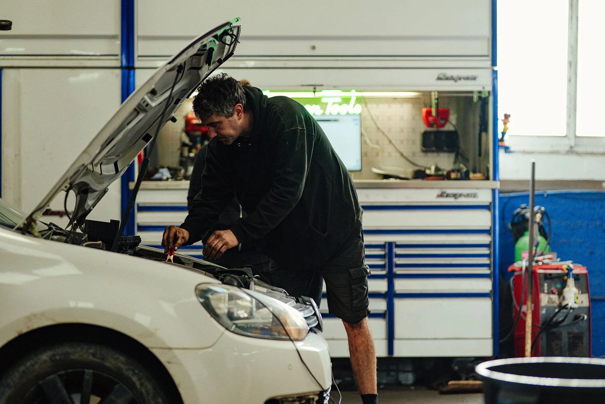 A man working under the hood of a white car in a garage with blue tool cabinets and a neon sign.