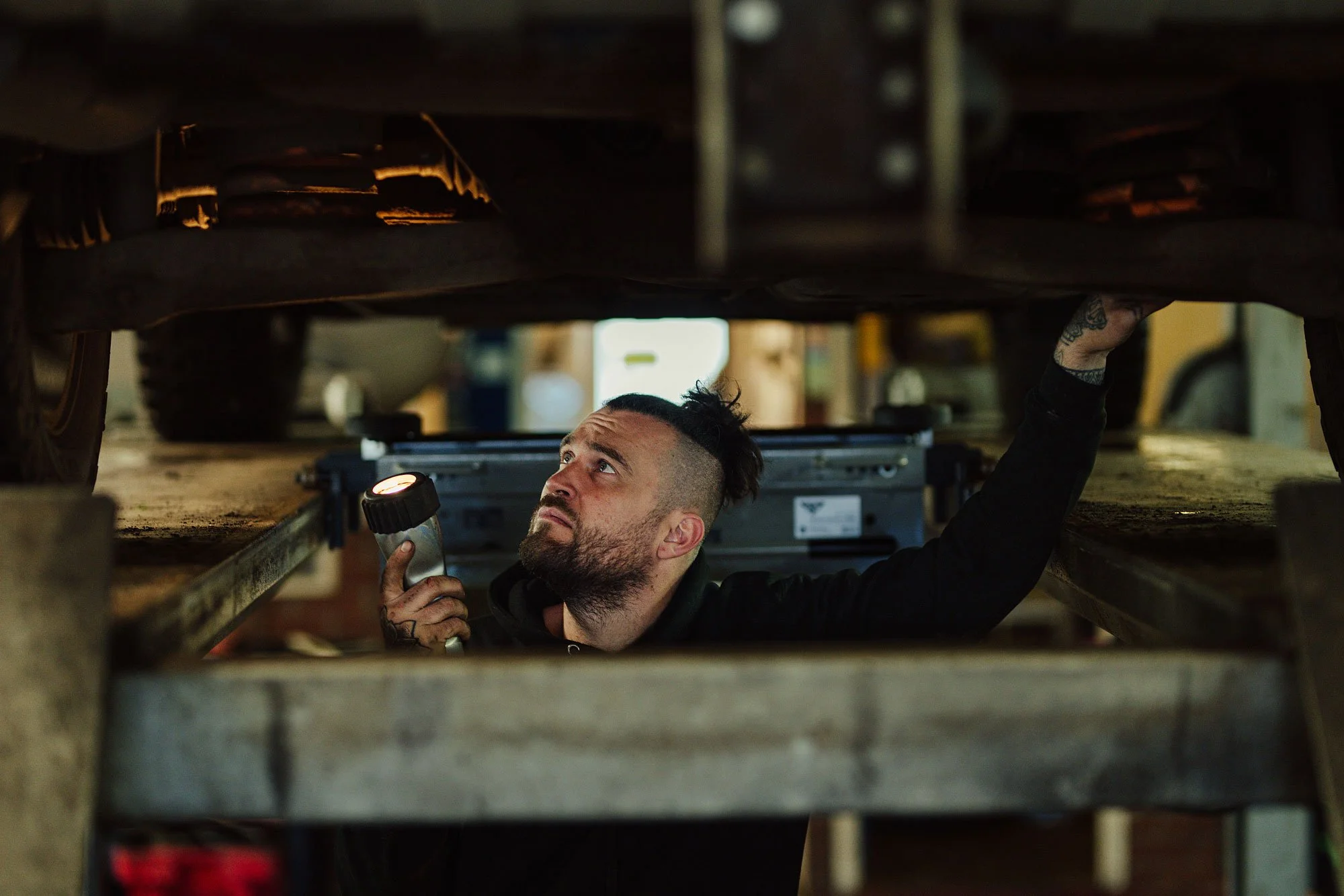 A man with a beard and tattoos working underneath a vehicle, holding a flashlight and examining the underside in a garage or workshop.