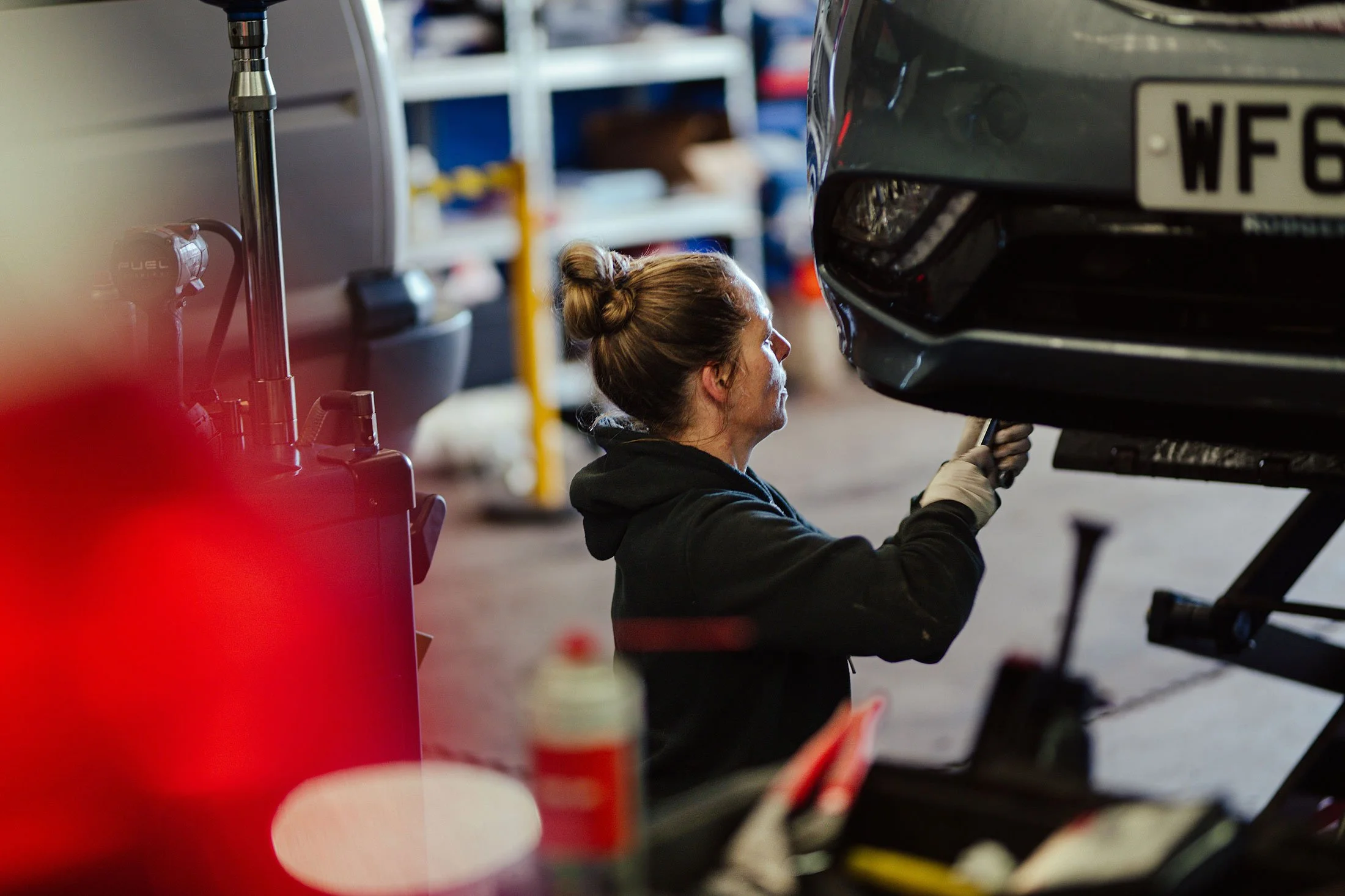 A woman in a black hoodie and gloves works underneath a car lifted on a hydraulic lift in a garage.