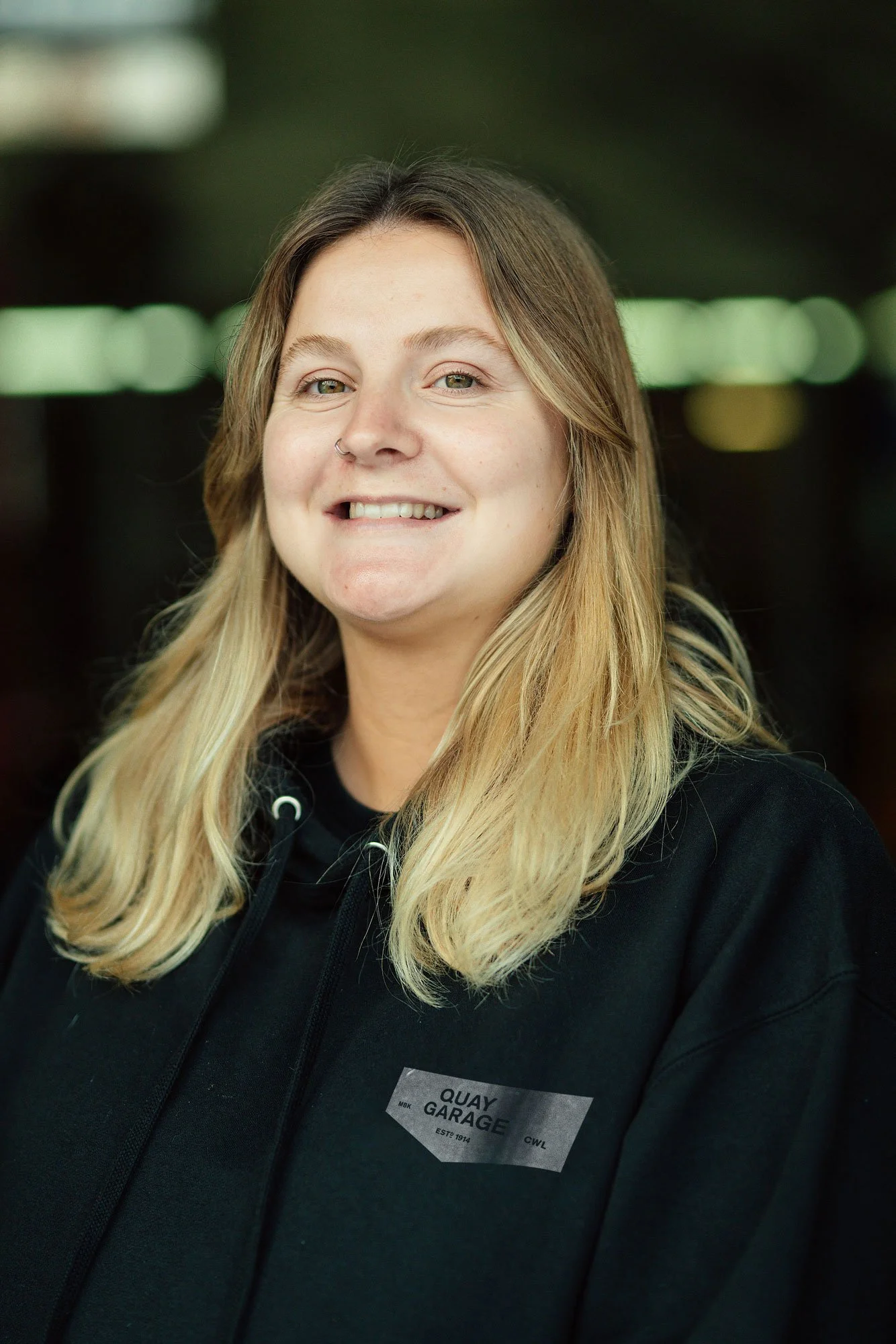 A woman with long blonde hair, smiling, wearing a black hoodie with a Quay Garage name tag in a dark indoor setting.