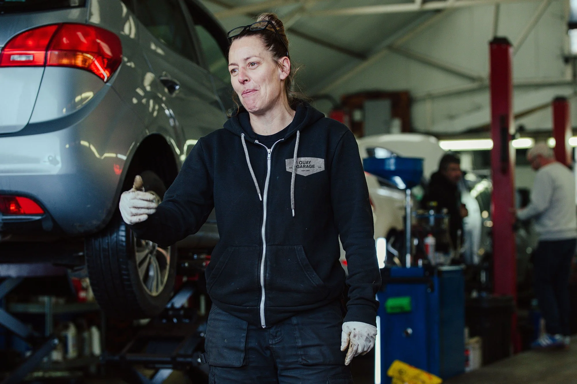 A woman in a black hoodie and gloves in an auto repair workshop, holding a small object, with a car on a lift in the background.