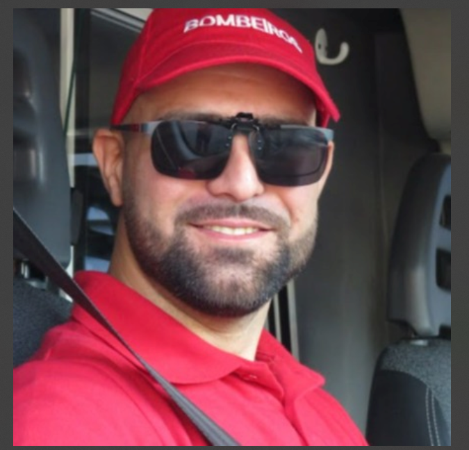 Homem com barba, óculos escuros, chapéu vermelho com a palavra "BOMBEIRO" e camiseta vermelha, sentado em um veículo de emergência.