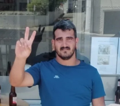Homem com barba, cabelo escuro, usando camiseta azul, fazendo sinal de paz com duas mãos, sentado em um ambiente interno com janela ao fundo.
