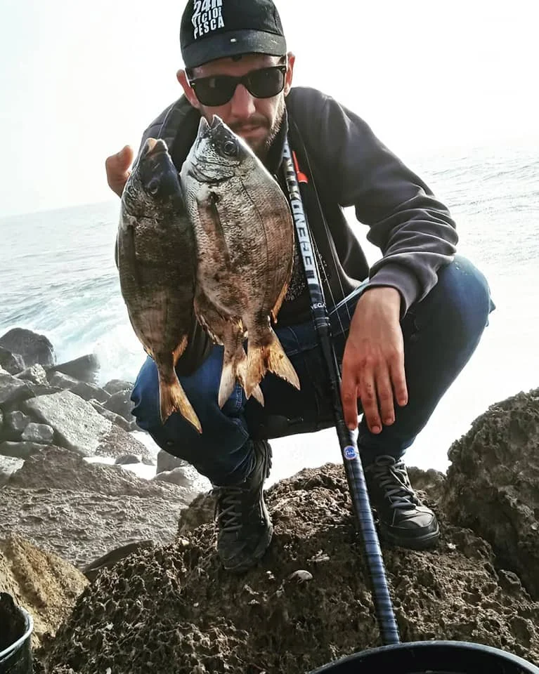 Homem com óculos escuros e boné negro segurando uma pescada e uma vara de pesca, na beira de rochas junto ao mar.