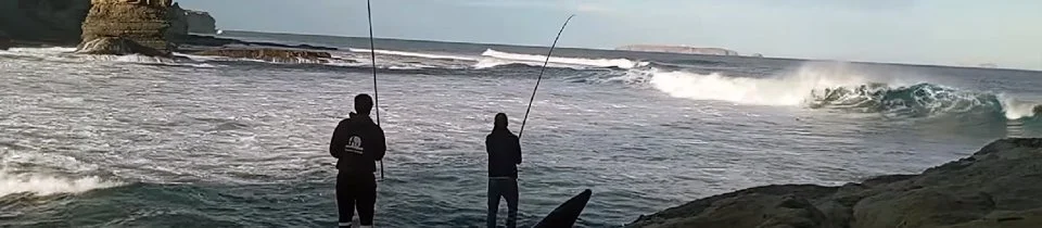 Duas pessoas pescando na praia, com ondas e uma falésia ao fundo.
