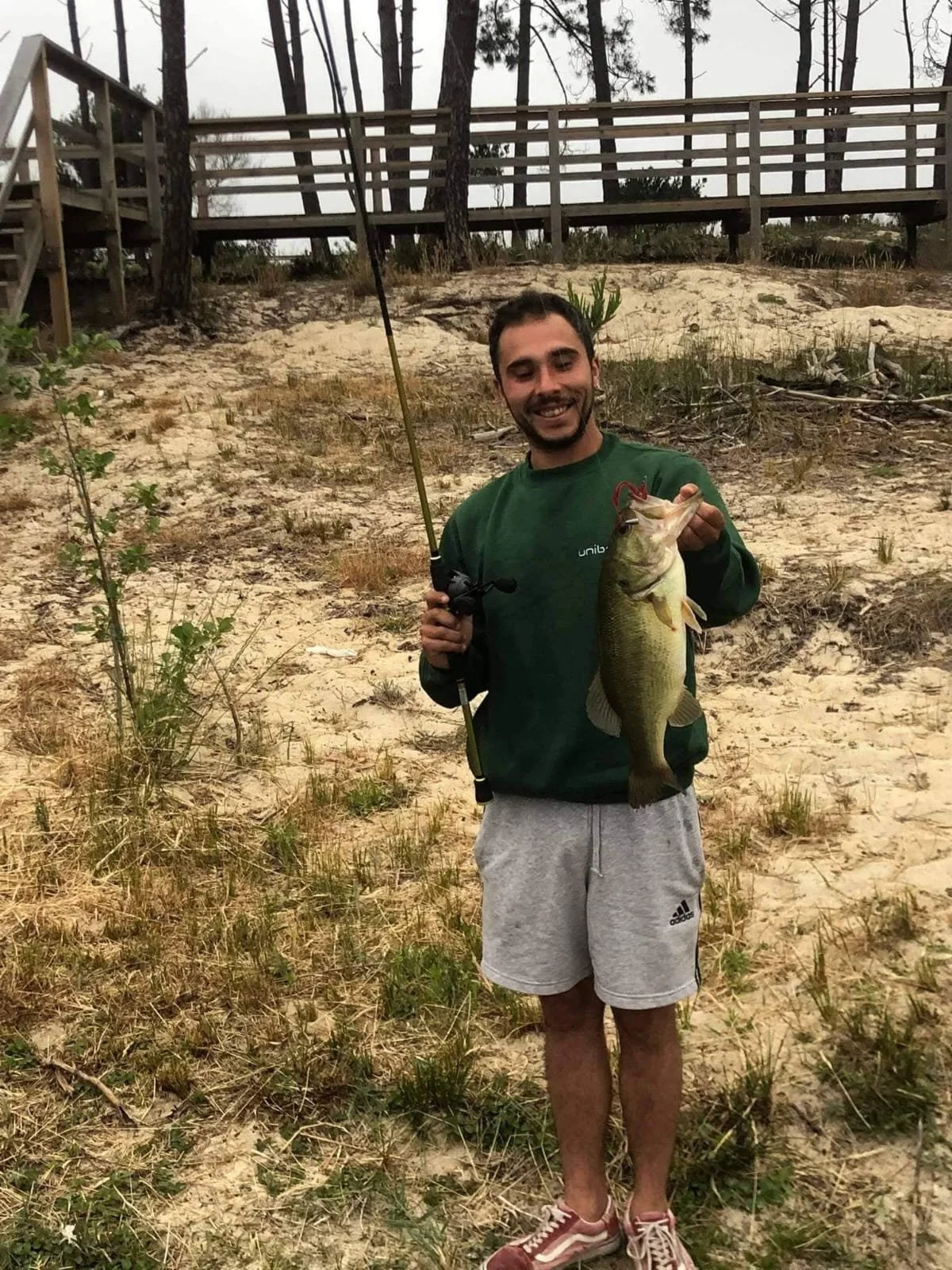 Homem sorridente segurando um peixe grande na mão direita, vestindo jaqueta verde, bermuda cinza e tênis, em uma área de praia com vegetação e uma ponte de madeira ao fundo.