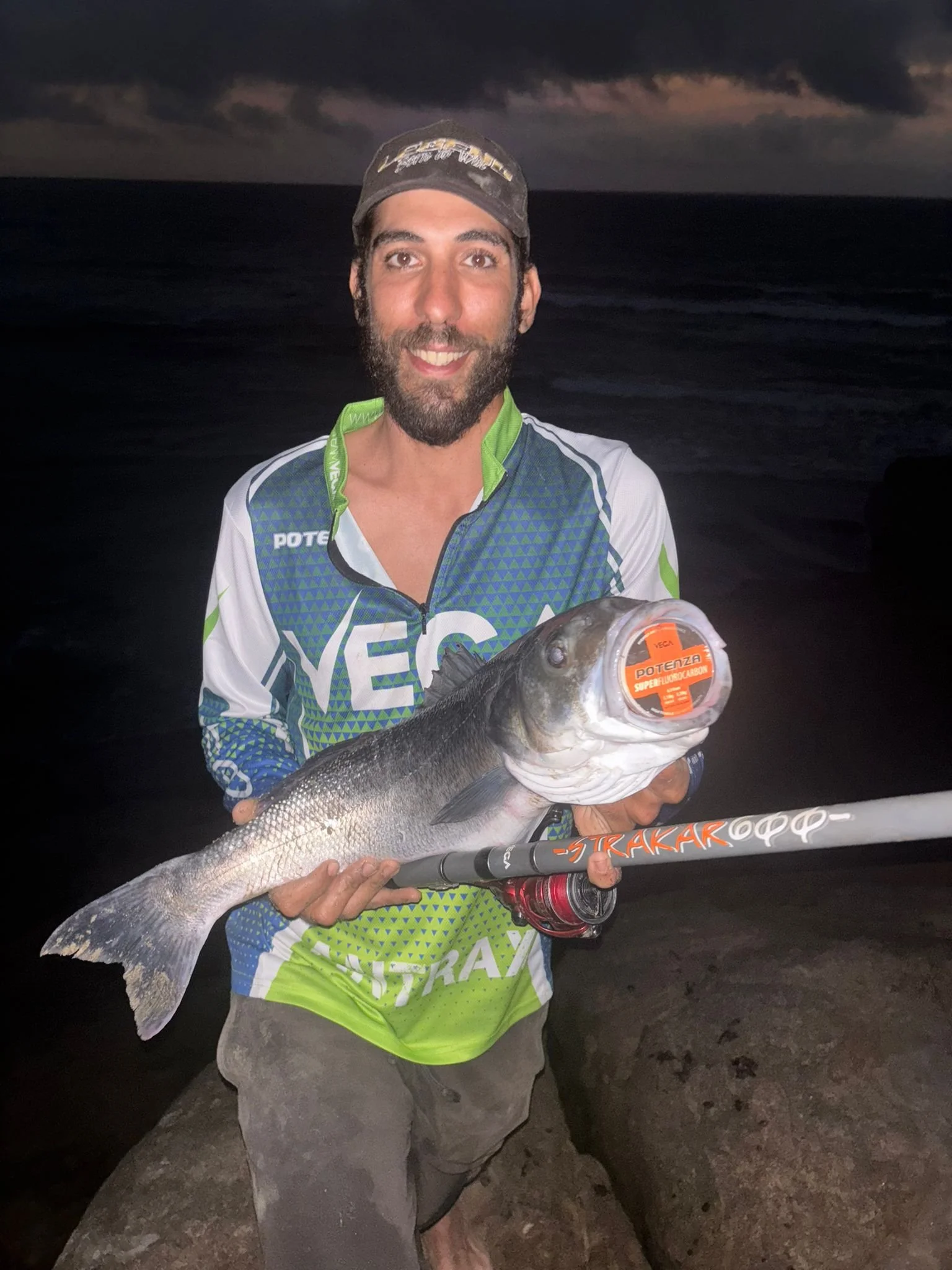 Homem sorridente segurando um peixe grande com uma vara de pescar, à noite na praia.