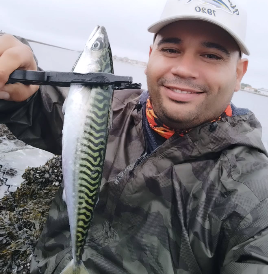 Homem segurando um peixe com uma pinça, ao ar livre perto da água, sorrindo para a câmera.