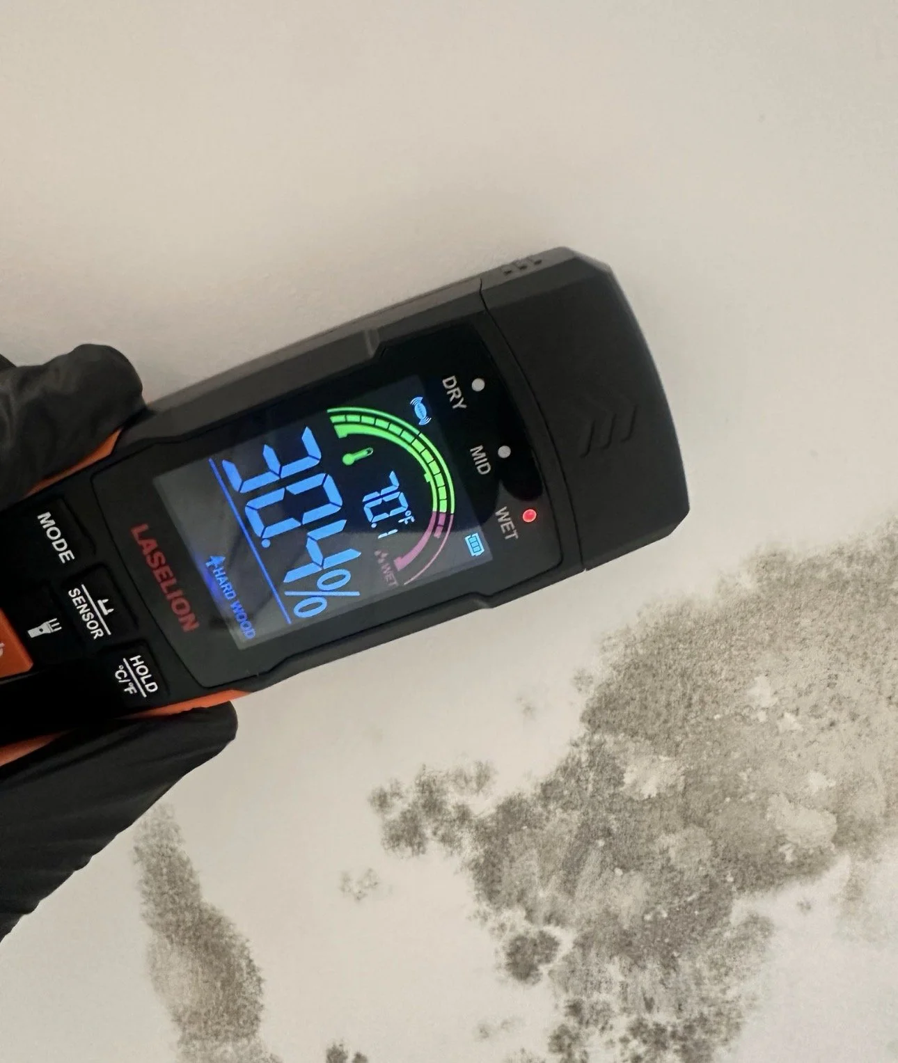 Infrared thermometer showing a temperature of 34.4°C, held over a white wall with dark mold patches.