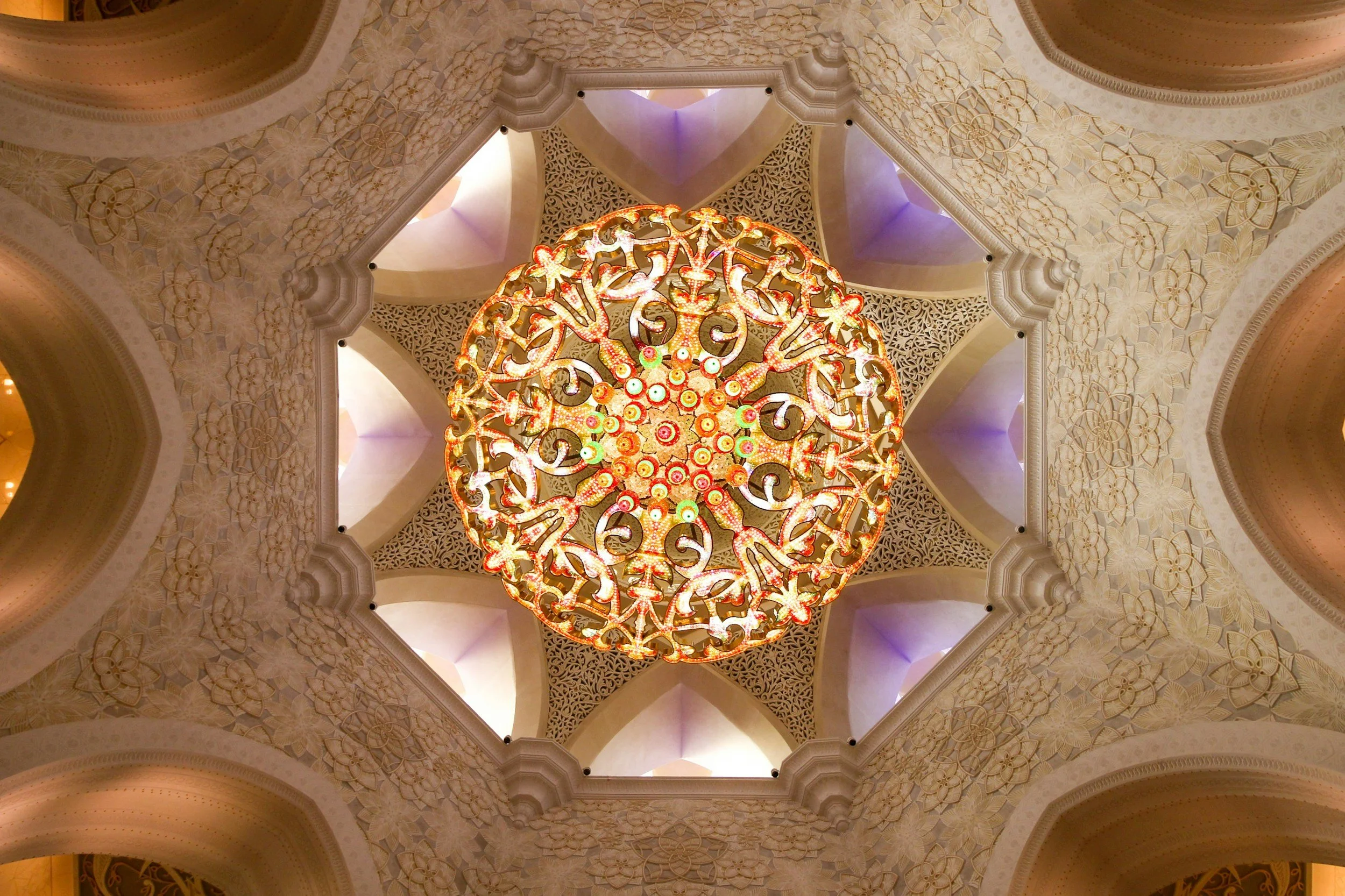 View of a large, ornate chandelier hanging from a decorated ceiling with multiple arches, intricate patterns, and soft lighting.