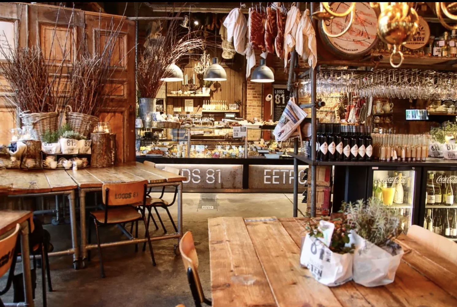 Interior of a cozy cafe with wooden tables and chairs, displaying decorative plants, hanging dried herbs, and a cozy counter area with various food items. The ambiance is warm and rustic with shelving, bottles, and glasses.