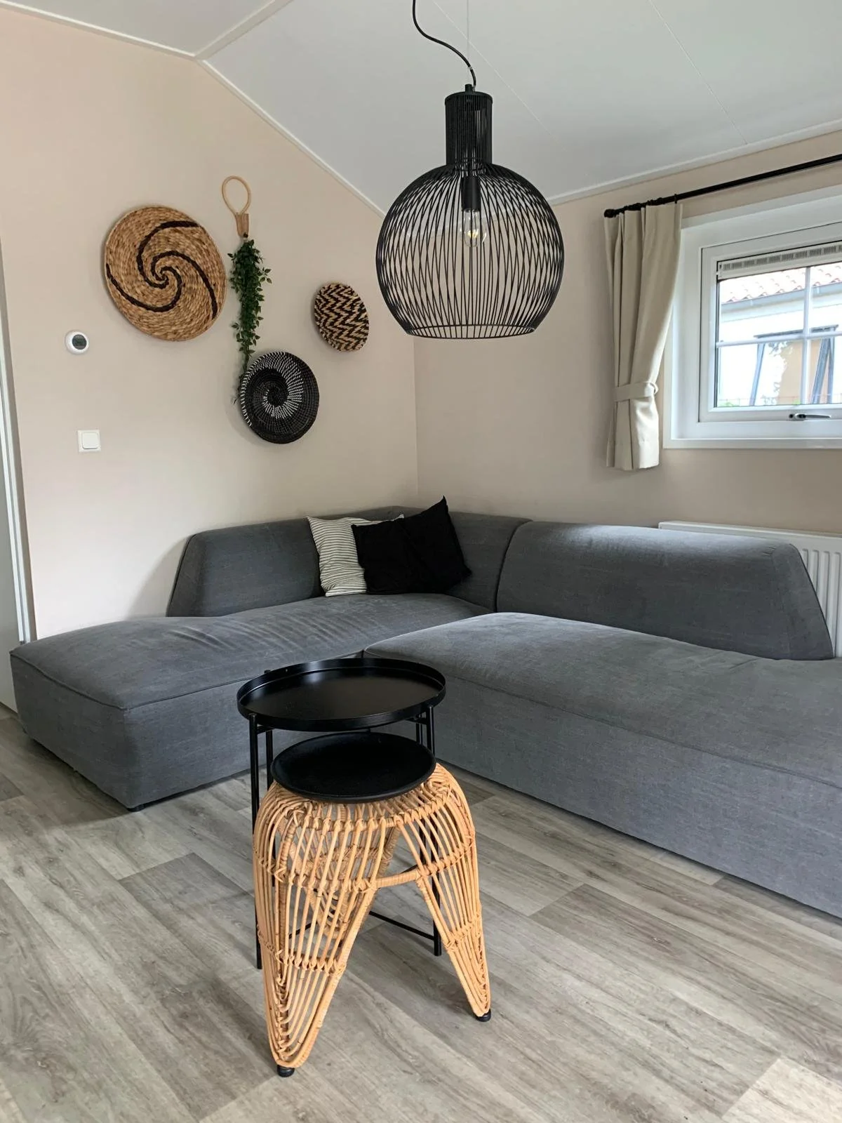 Living room with a gray sectional sofa, black and white pillows, a black wire pendant light, wall decor with woven baskets, a window with white curtains, and a black and wicker side table.