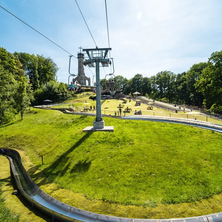 A water park with a large water slide and a ski lift, surrounded by green grass and trees, with people sitting on benches and walking around.