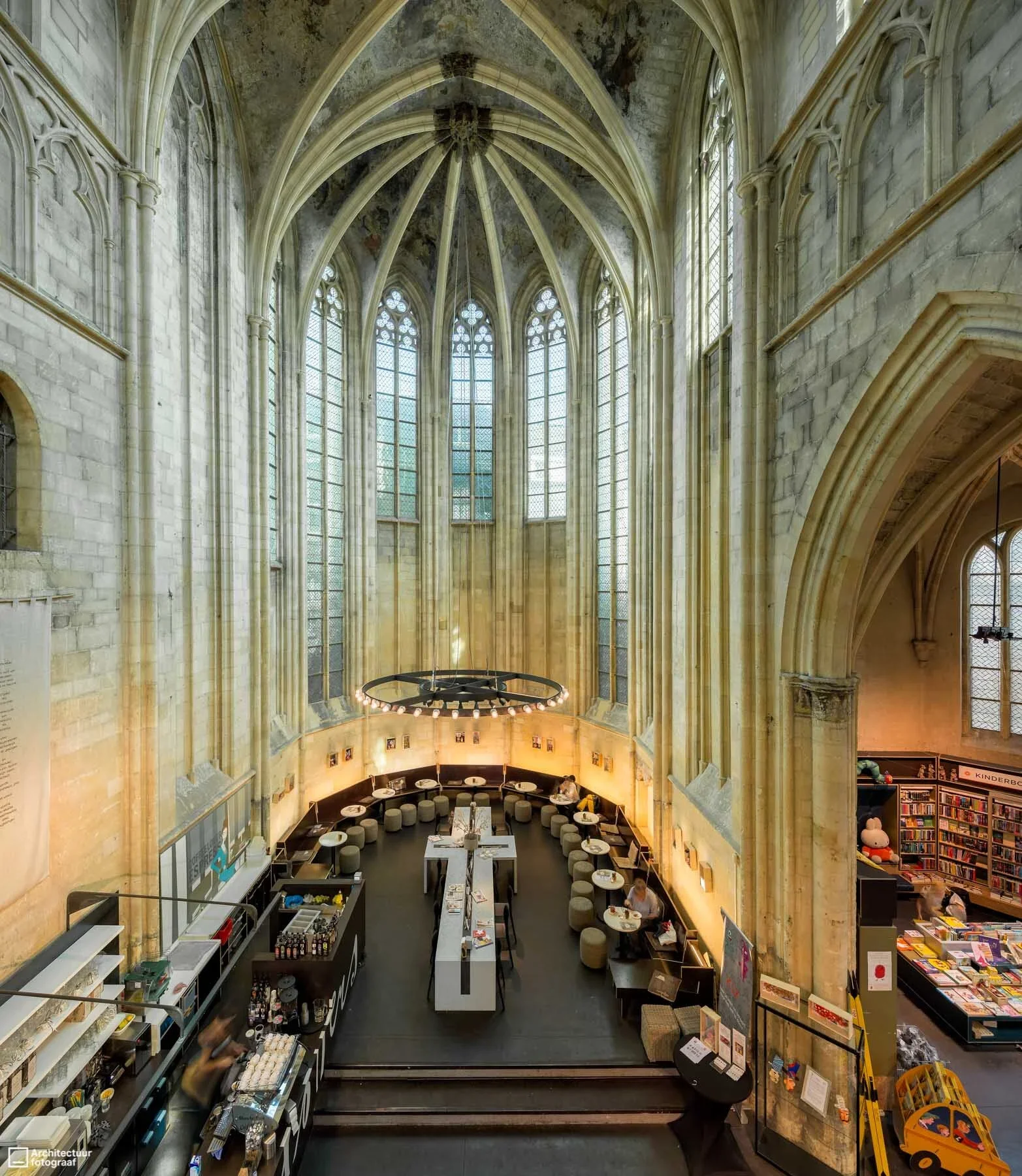 Interior view of a large, historic church converted into a modern cafe or bookstore, with high stained glass windows, stone walls, and a circular seating area with small tables and stools, lit by a contemporary circular chandelier.