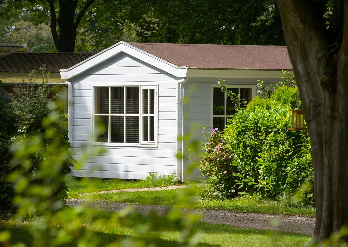 A white house with a brown roof partially obscured by green bushes and trees, featuring large multi-pane windows.