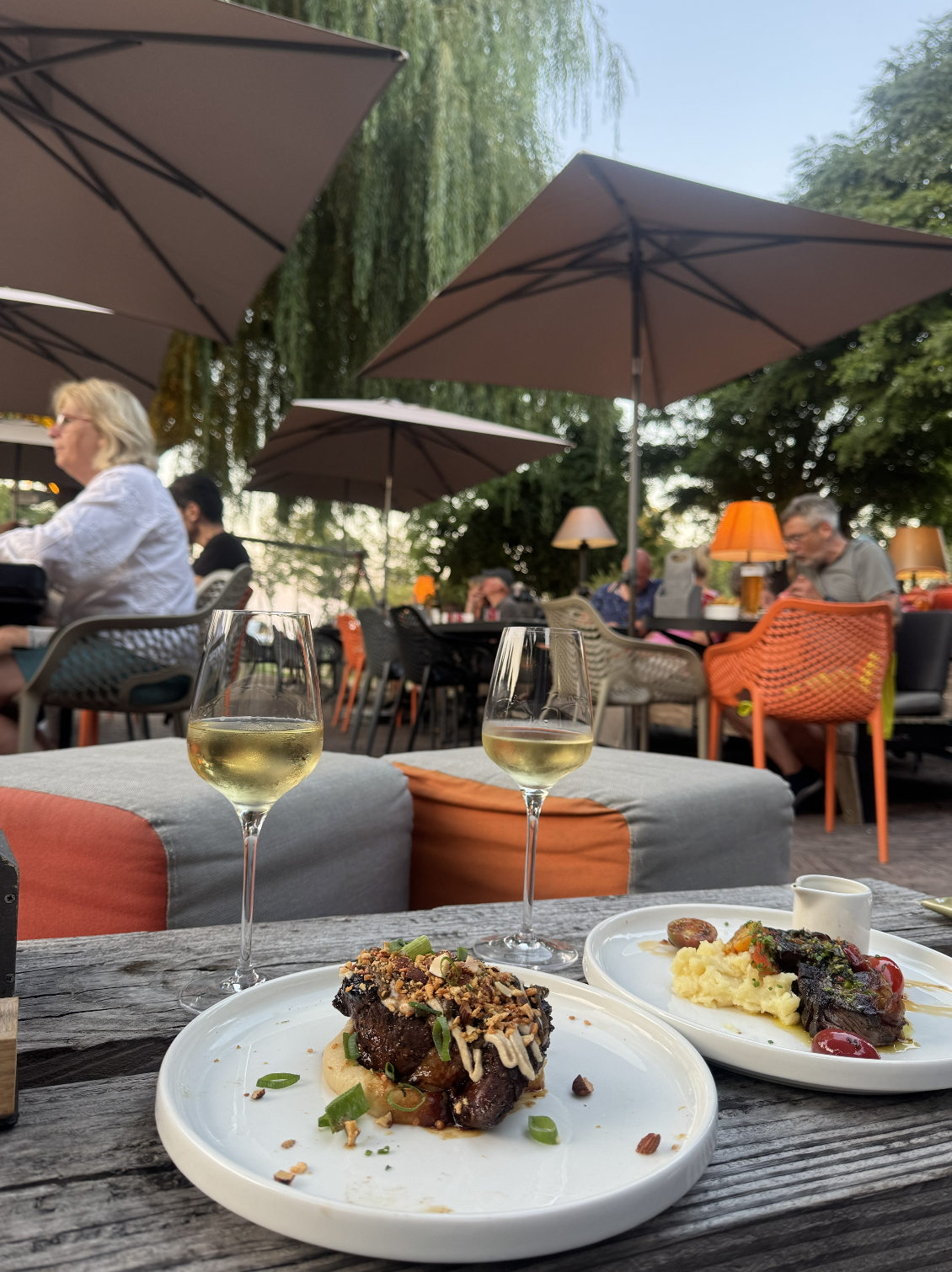 Two glasses of white wine on a wooden table in an outdoor restaurant patio, with dishes of meat and mashed potatoes topped with vegetables. There are patio furniture and umbrellas, and people dining in the background.