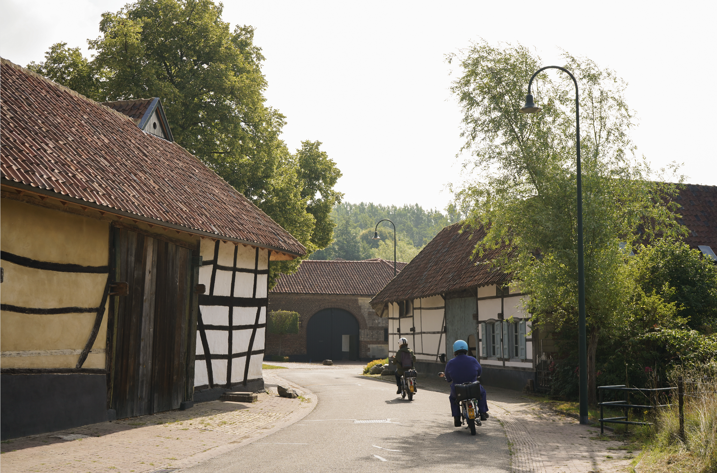 Two people riding electric bikes down a narrow street in a rural village with traditional cottages, trees, and street lamps.
