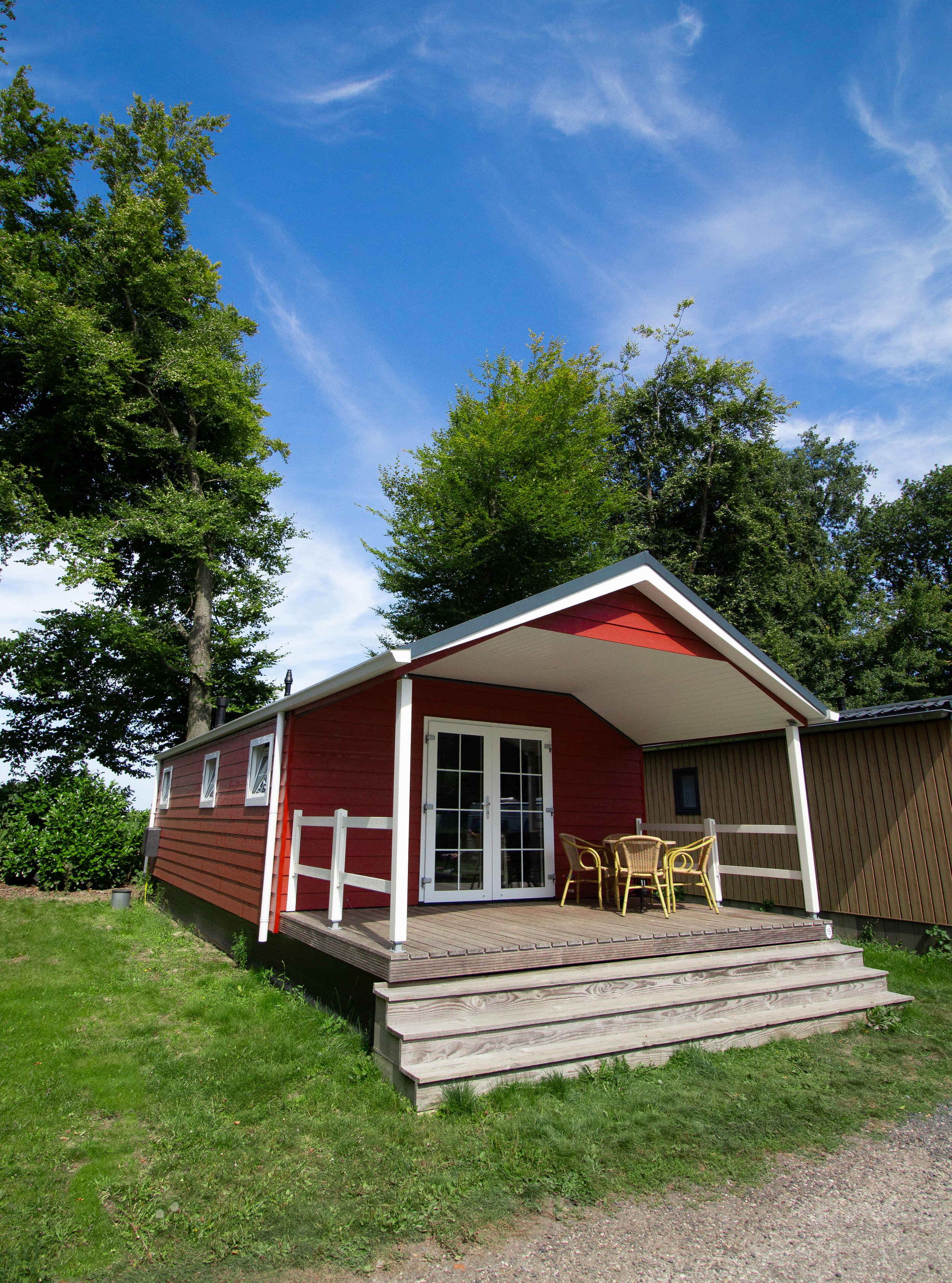 A small red wooden house with a covered porch and outdoor dining table with chairs, set on a grassy area under a blue sky with clouds, surrounded by tall trees.
