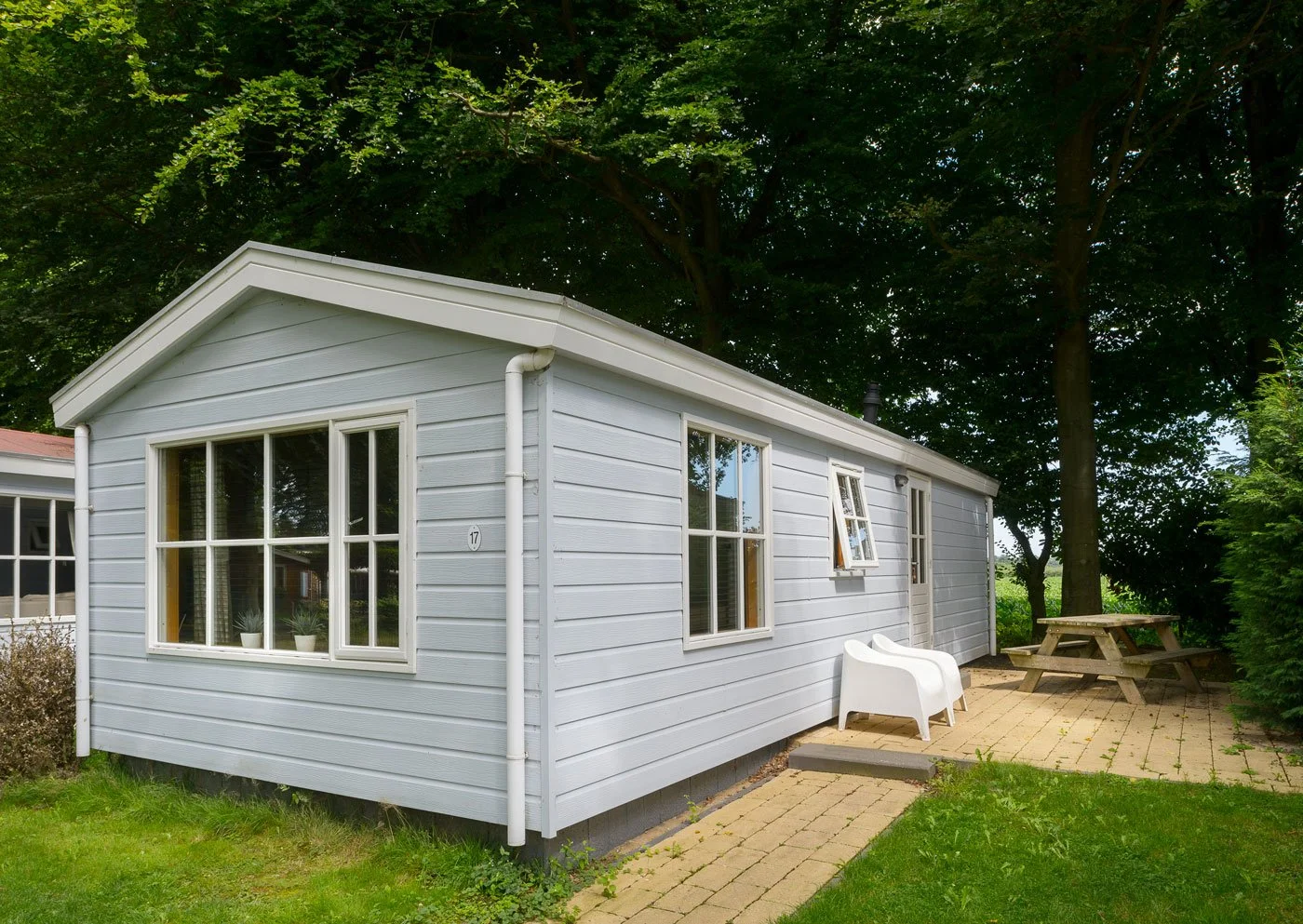 A light gray house with white trim, a gabled roof, and a small brick patio with outdoor furniture and a picnic table, surrounded by green trees and grass.