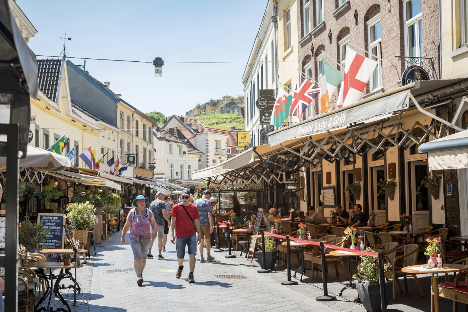 People walking along a lively outdoor street with cafes and restaurants, flags from different countries hanging above, and a hill with buildings in the background on a sunny day.