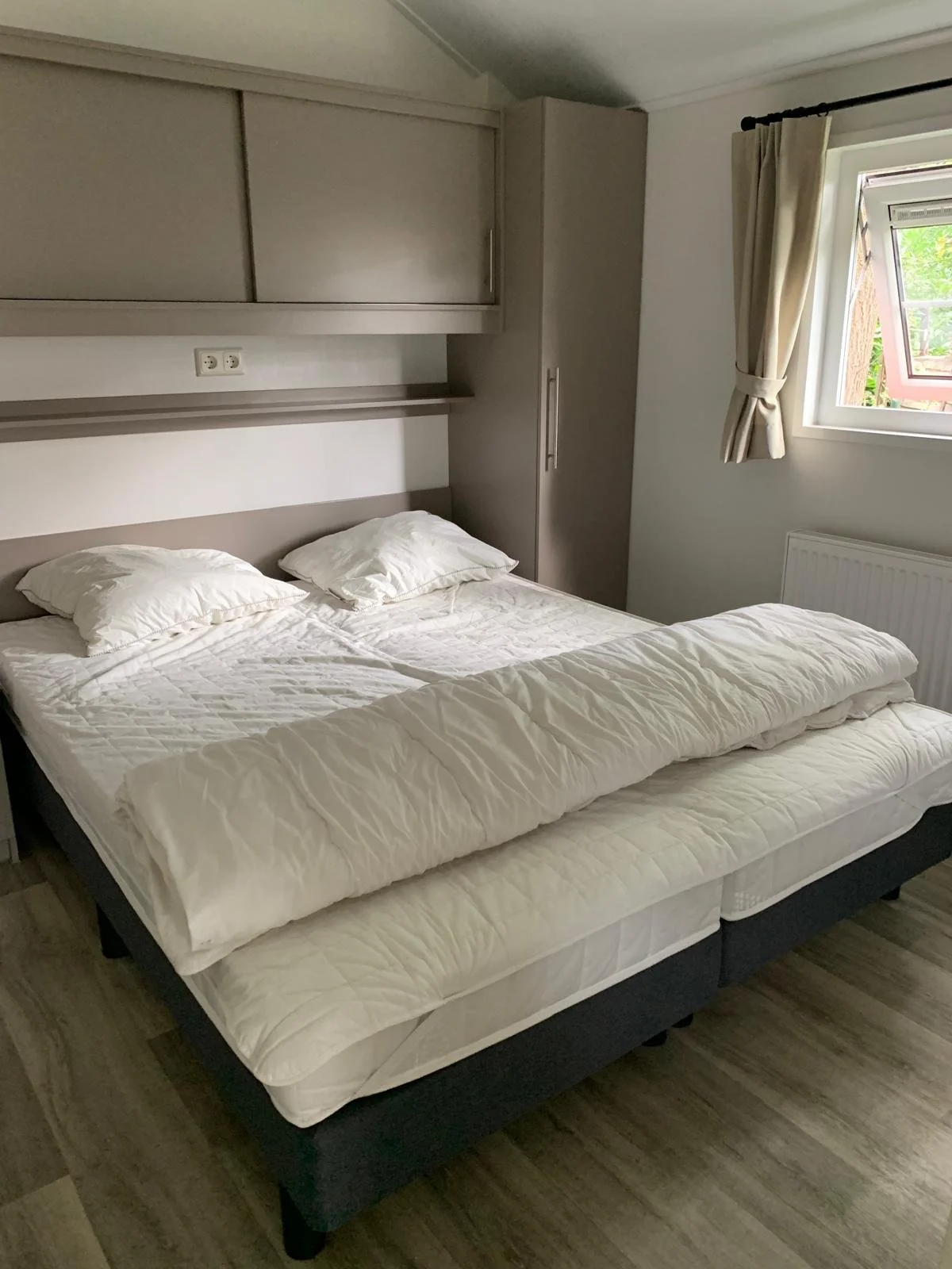 A neatly made bed with white bedding and two pillows in a bedroom with a window showing greenery outside.