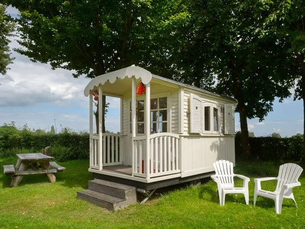 A small white playhouse with a porch, red lanterns, and shutters, located in a grassy yard under a large tree, with two white lawn chairs and a picnic table nearby.