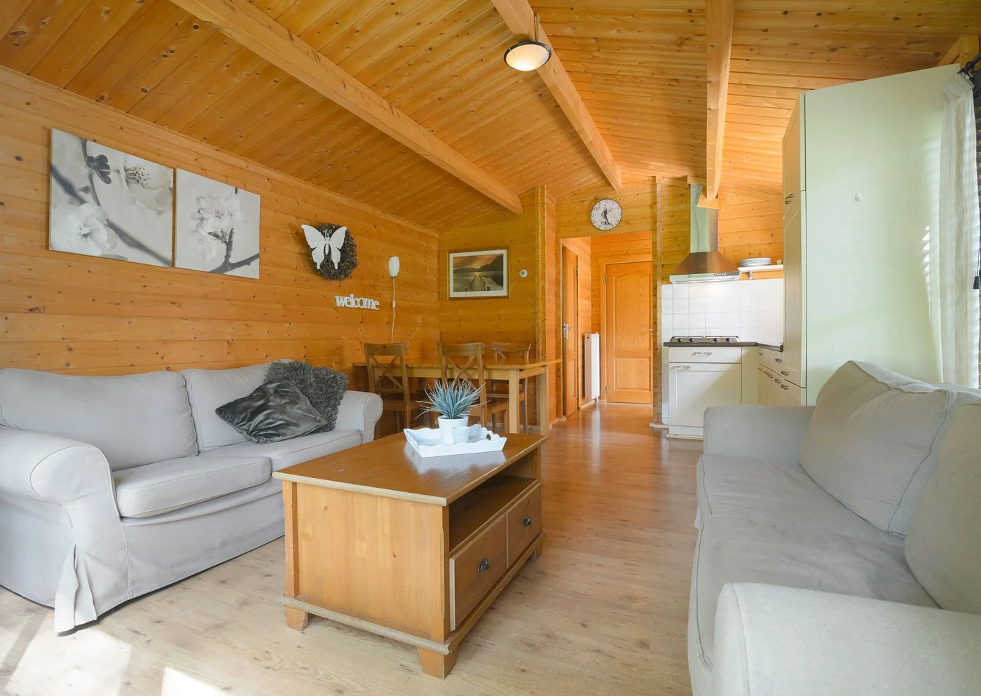 Living room with wood-paneled walls and ceiling, two beige sofas, a wooden coffee table with a potted plant, a small dining table with chairs, and a visible kitchen area with white cabinets and a stove. Decor includes wall art, a clock, a butterfly o