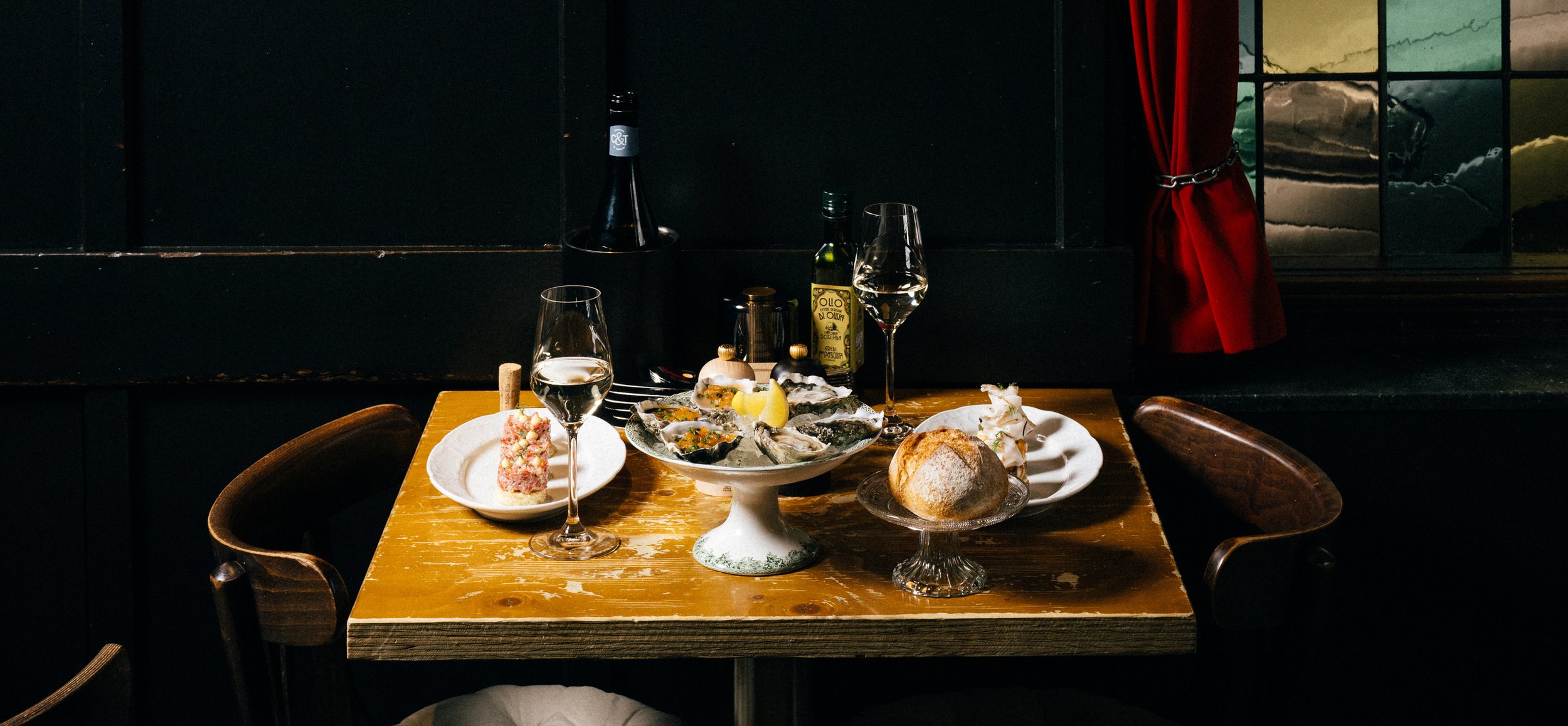 A rustic wooden table set with various foods, including oysters on a pedestal, a bread roll, a dessert, and a glass of white wine. There are bottles of olive oil and a wine bottle in the background. The setting appears cozy with dark walls and a red curtain.