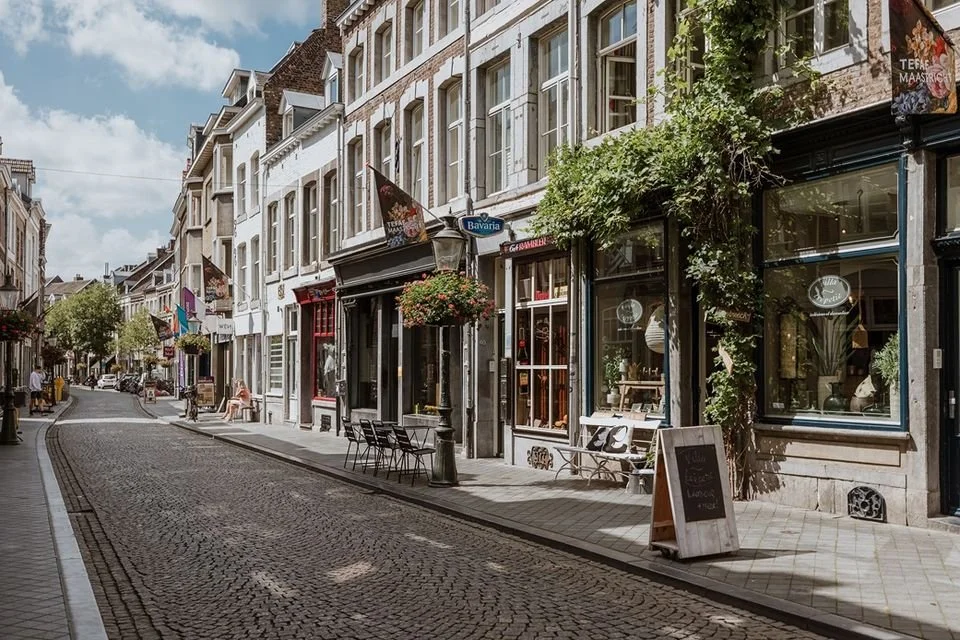 A quiet cobblestone street in a European city with historic buildings, storefronts, and outdoor seating. There are hanging flower baskets, a bench, and people walking or sitting.