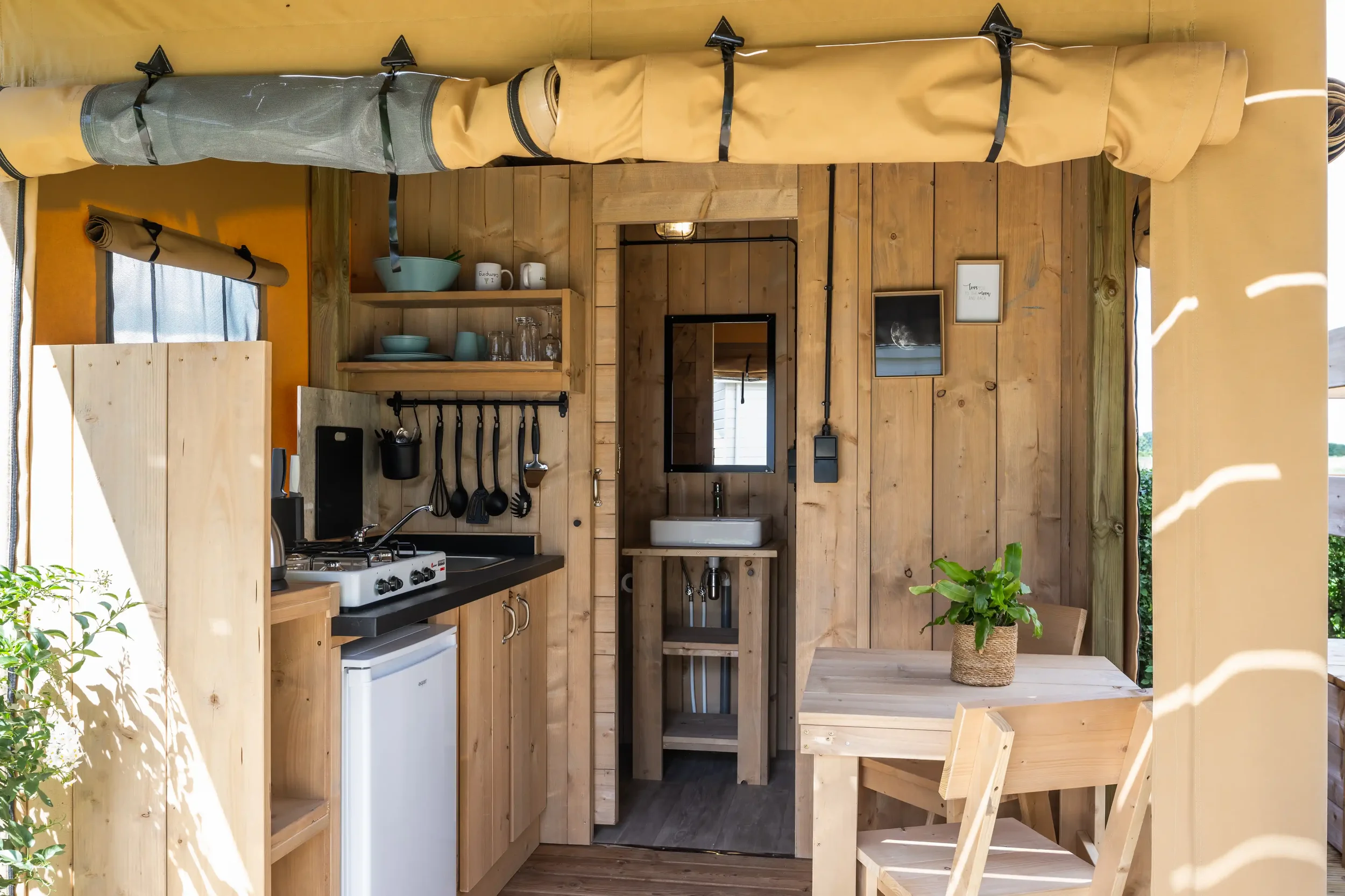 Interior of a tiny wooden kitchen with a small table, a potted plant, a sink, shelves with dishes, and hanging utensils.
