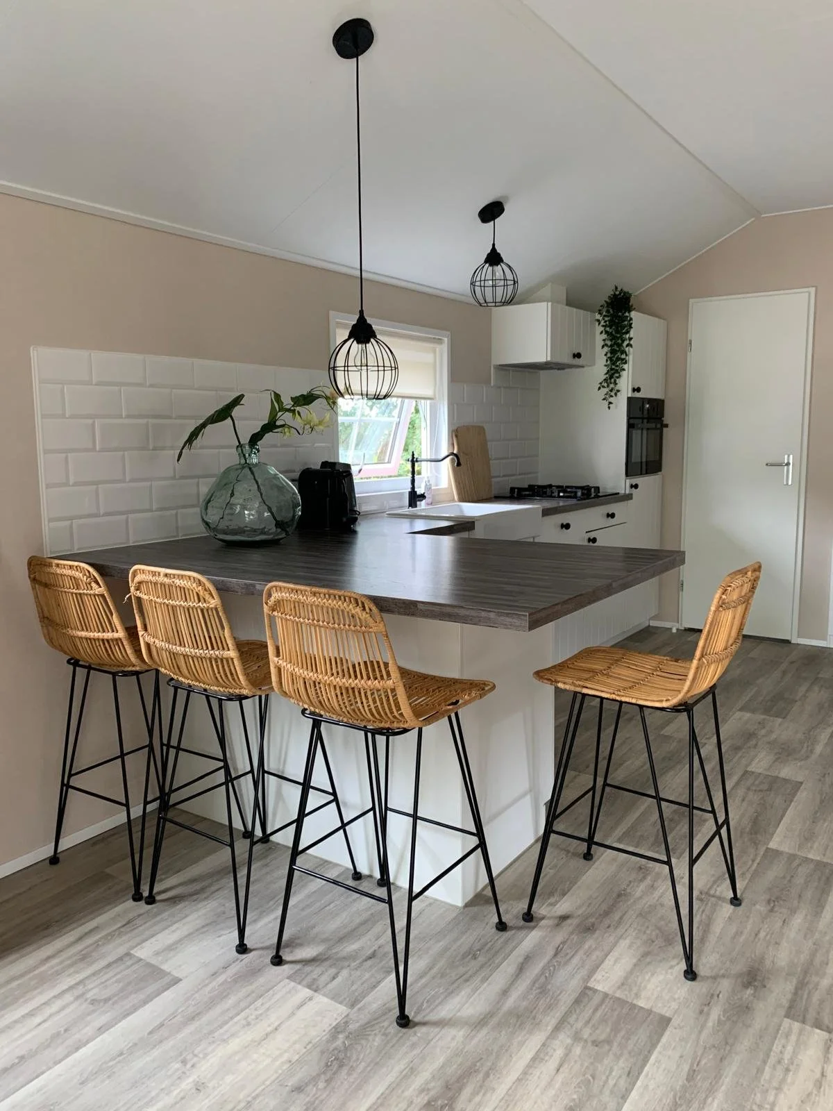 A modern kitchen with a dark wood countertop island, four rattan bar stools, a white subway tile backsplash, white cabinets, a window, a black faucet, a small potted plant, and hanging black metal pendant lights.