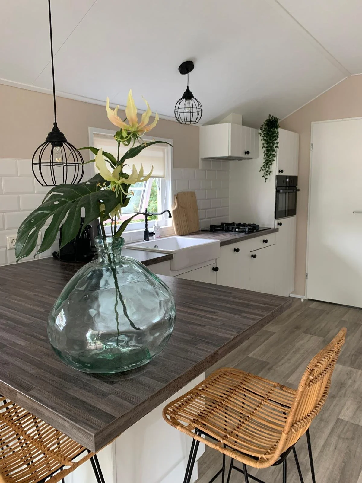 Modern kitchen interior with a dark wooden kitchen island, clear glass vase with green and white flowers, rattan bar stool, white cabinets, and black pendant lights.