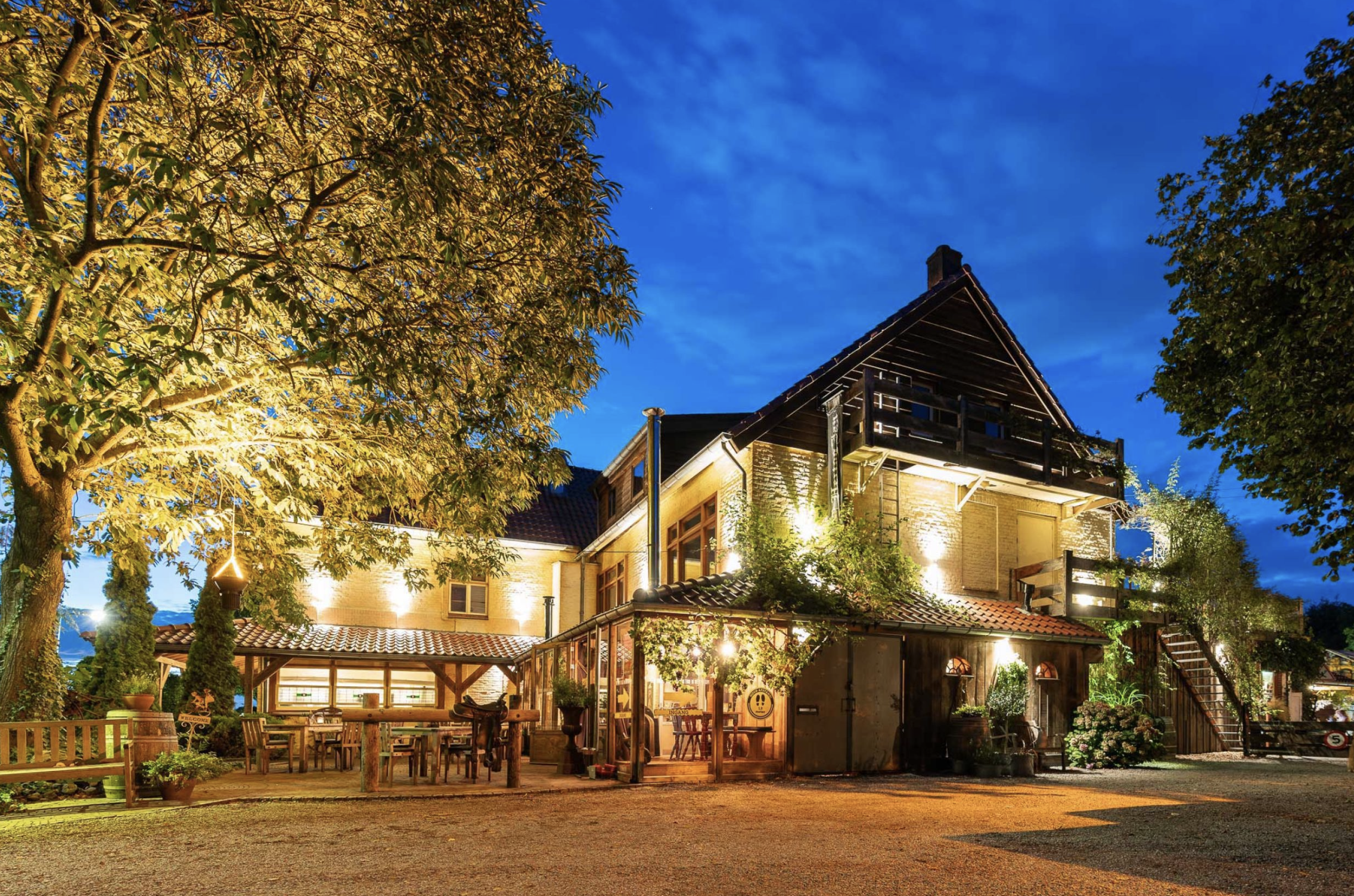 A warmly lit house with outdoor dining area, surrounded by trees and plants, under a blue evening sky.