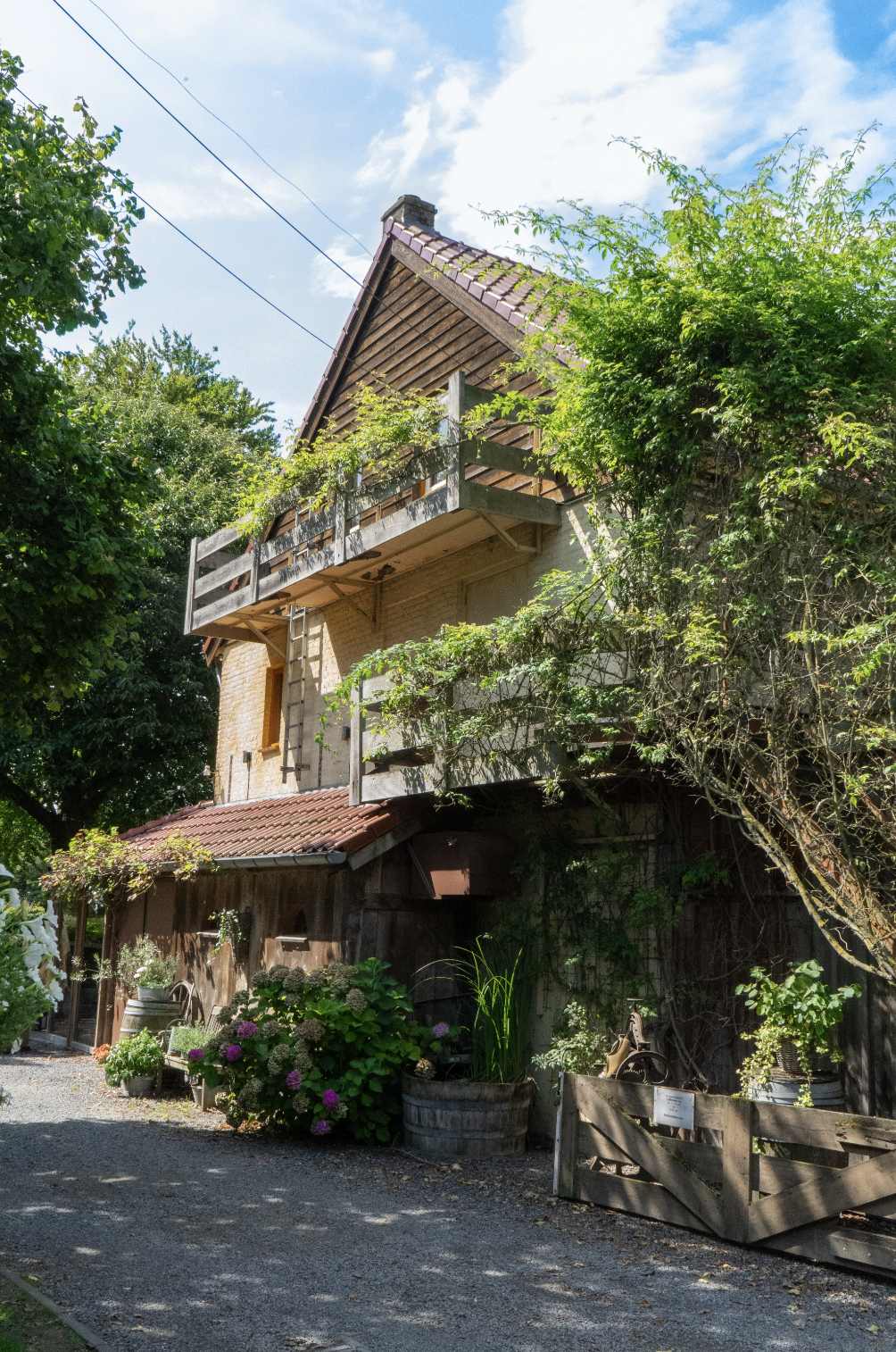 A rustic, multi-level house with a sloped roof and a wooden balcony, surrounded by lush greenery and colorful flowers, under a partly cloudy sky.