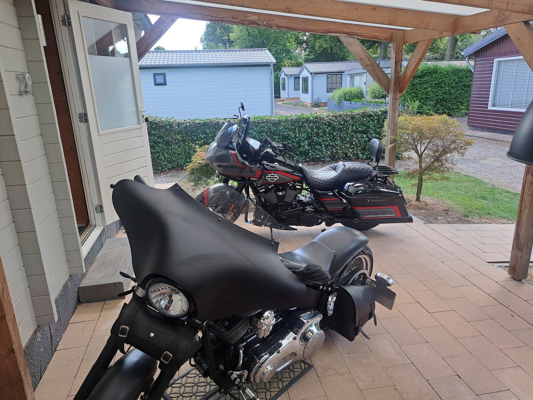 Twee Harley Davidson motorfietsen geparkeerd op een terras onder een houten afdak, met een witte deur en een achtergrond van kleine huisjes en groene struiken.