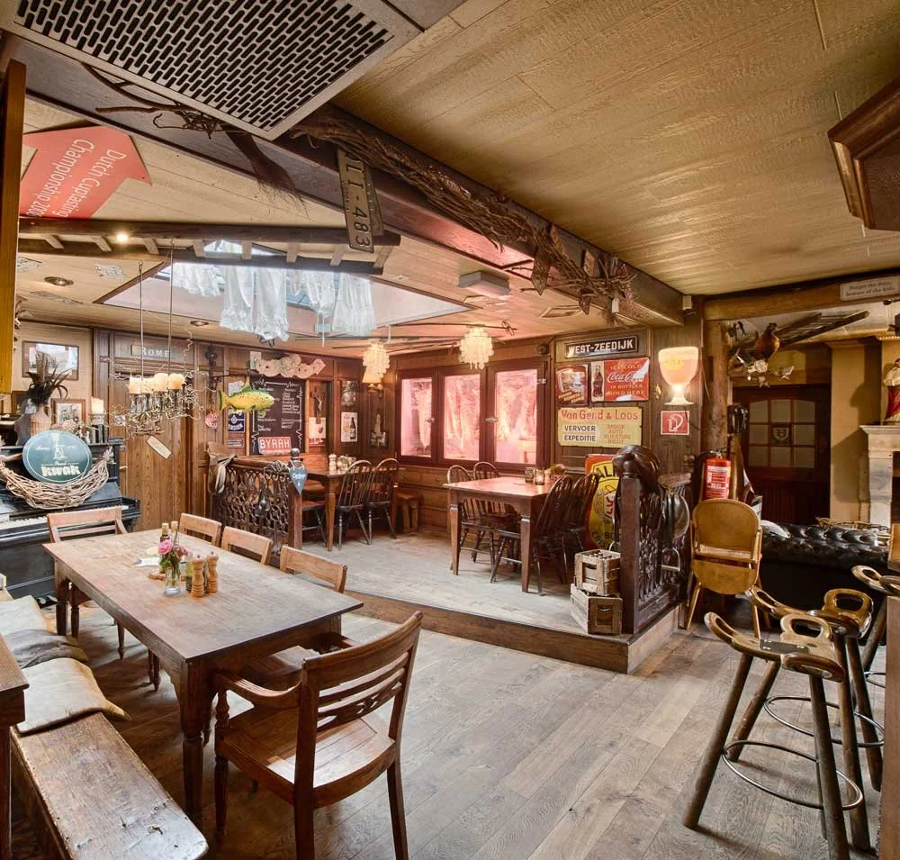 Interior of a cozy, vintage-style restaurant or pub with wooden furniture, signs, and decorative lighting, featuring a raised platform area and a mix of chairs and tables.