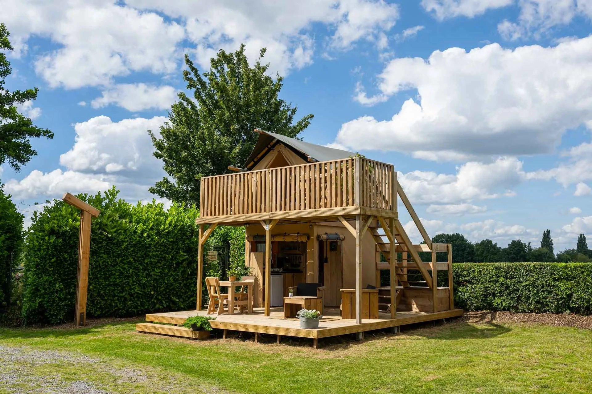 A two-story wooden playhouse with an upper deck and stairs, situated on a grassy lawn under a blue sky with clouds. The playhouse has a shaded outdoor area with furniture and is surrounded by green bushes.