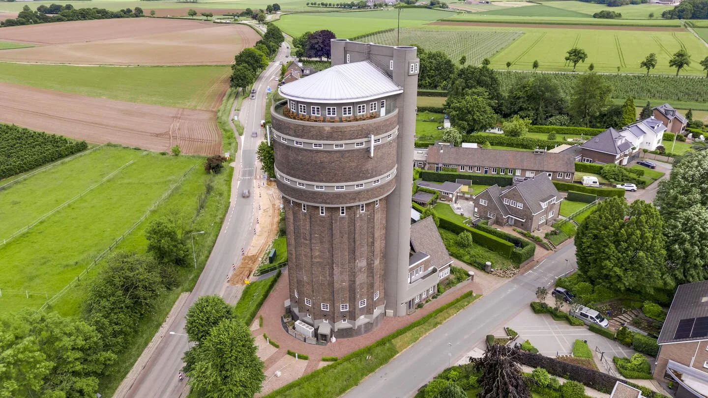 An aerial view of a tall, cylindrical water tower with a brick exterior and a rounded top, surrounded by roads, trees, and residential houses, with farmland in the background.