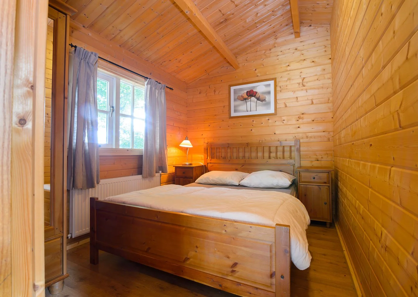 A cozy bedroom with wooden walls, a bed with white sheets and pillows, wooden nightstands, a small lamp, a window with curtains, and a framed picture of flowers above the bed.