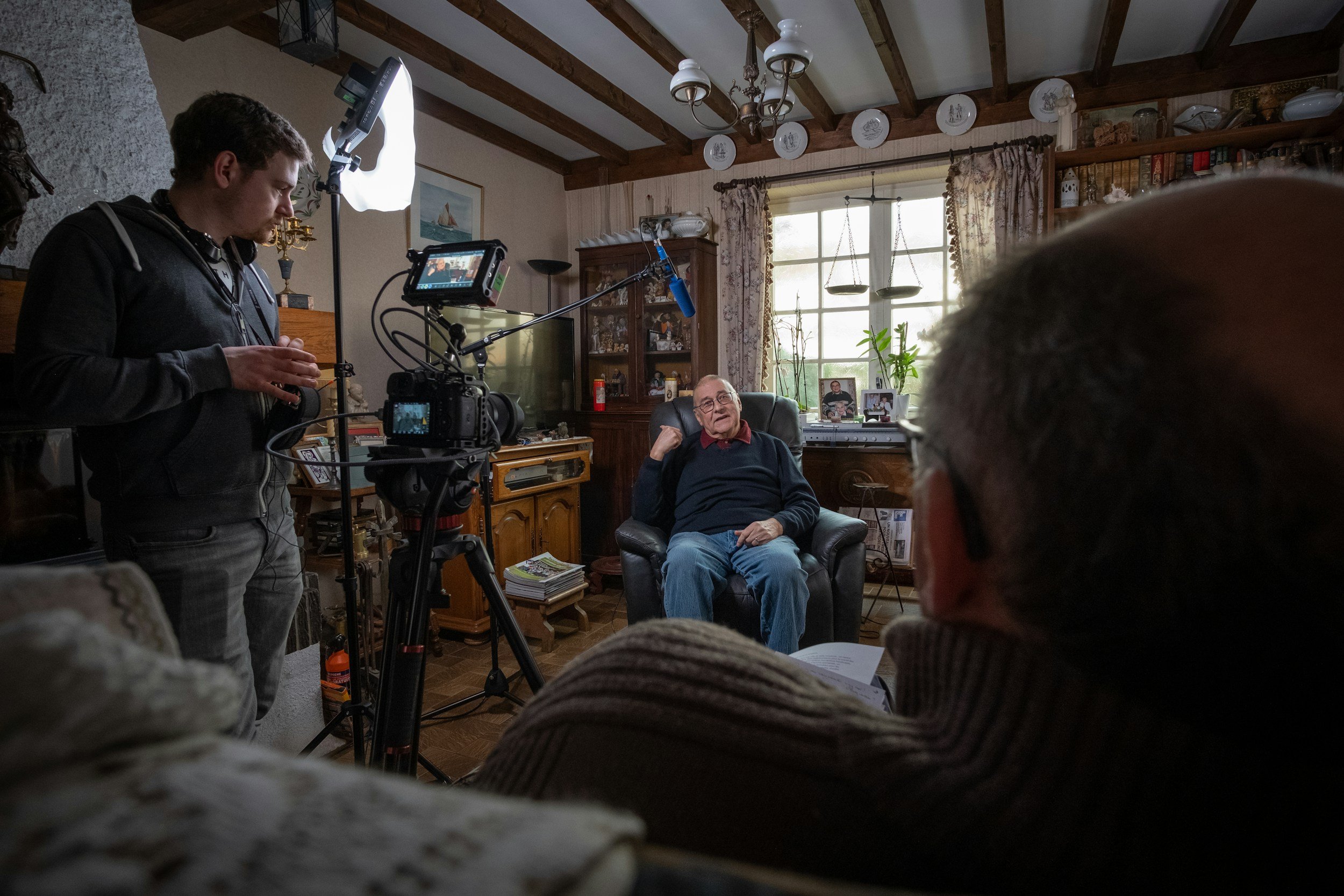 Filming a scene in a cozy living room with a camera operator on the left, an elderly man in a black sweater sitting in an armchair in the center, and another person in the foreground with glasses, all participating in an interview or recording session.