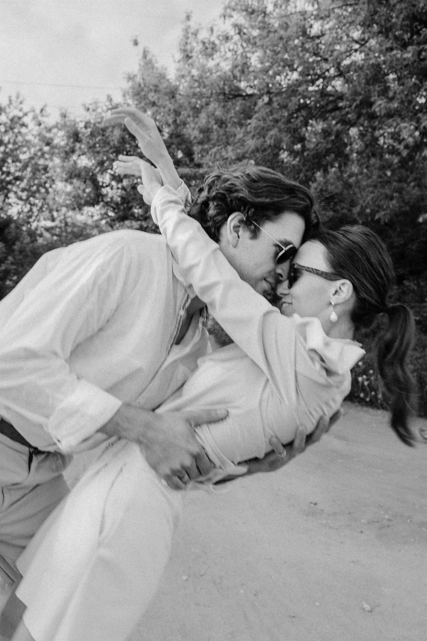 A man and woman embrace each other closely outdoors, both wearing sunglasses, with trees in the background.